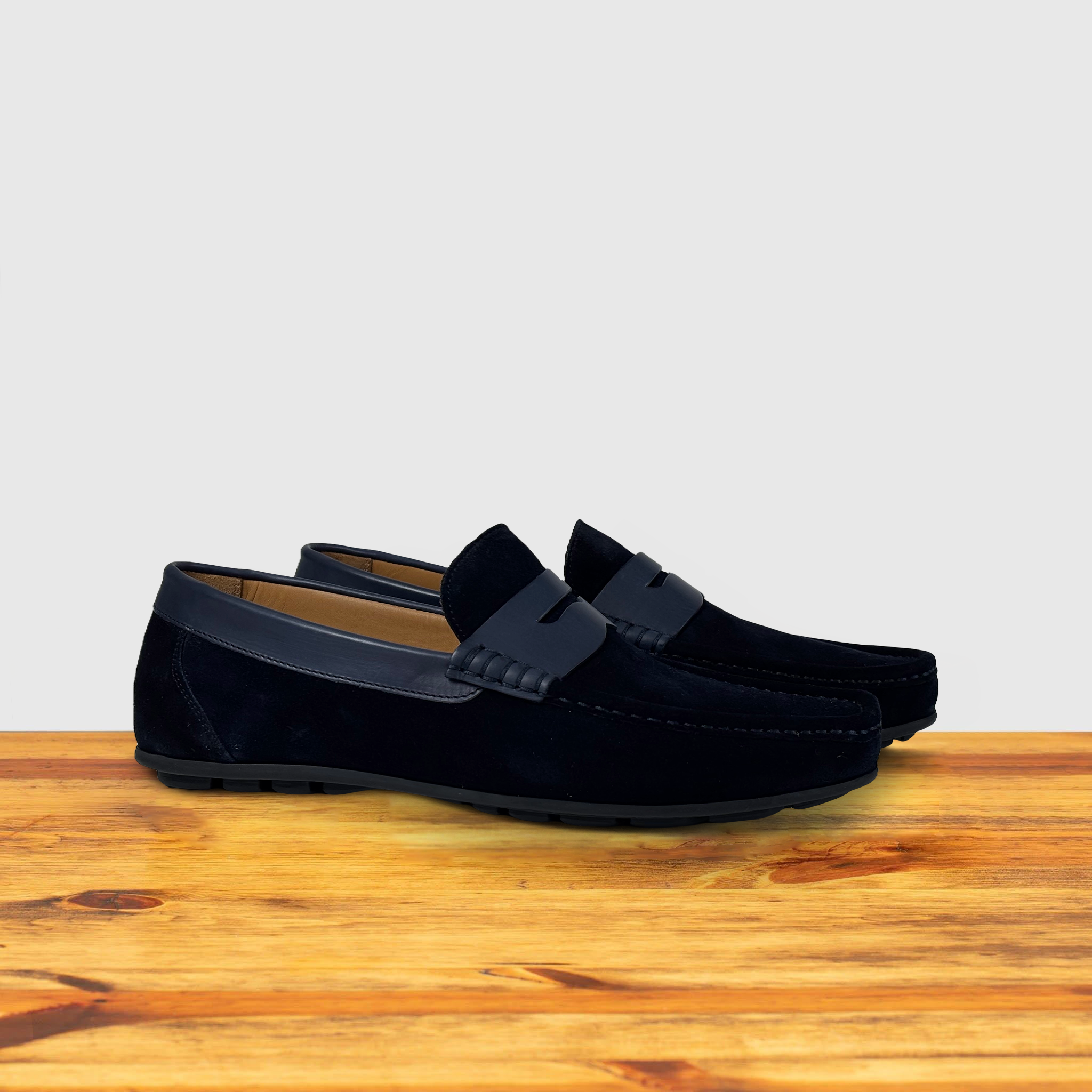 Pair of navy blue suede penny loafer with a strap, displayed on a wooden surface.