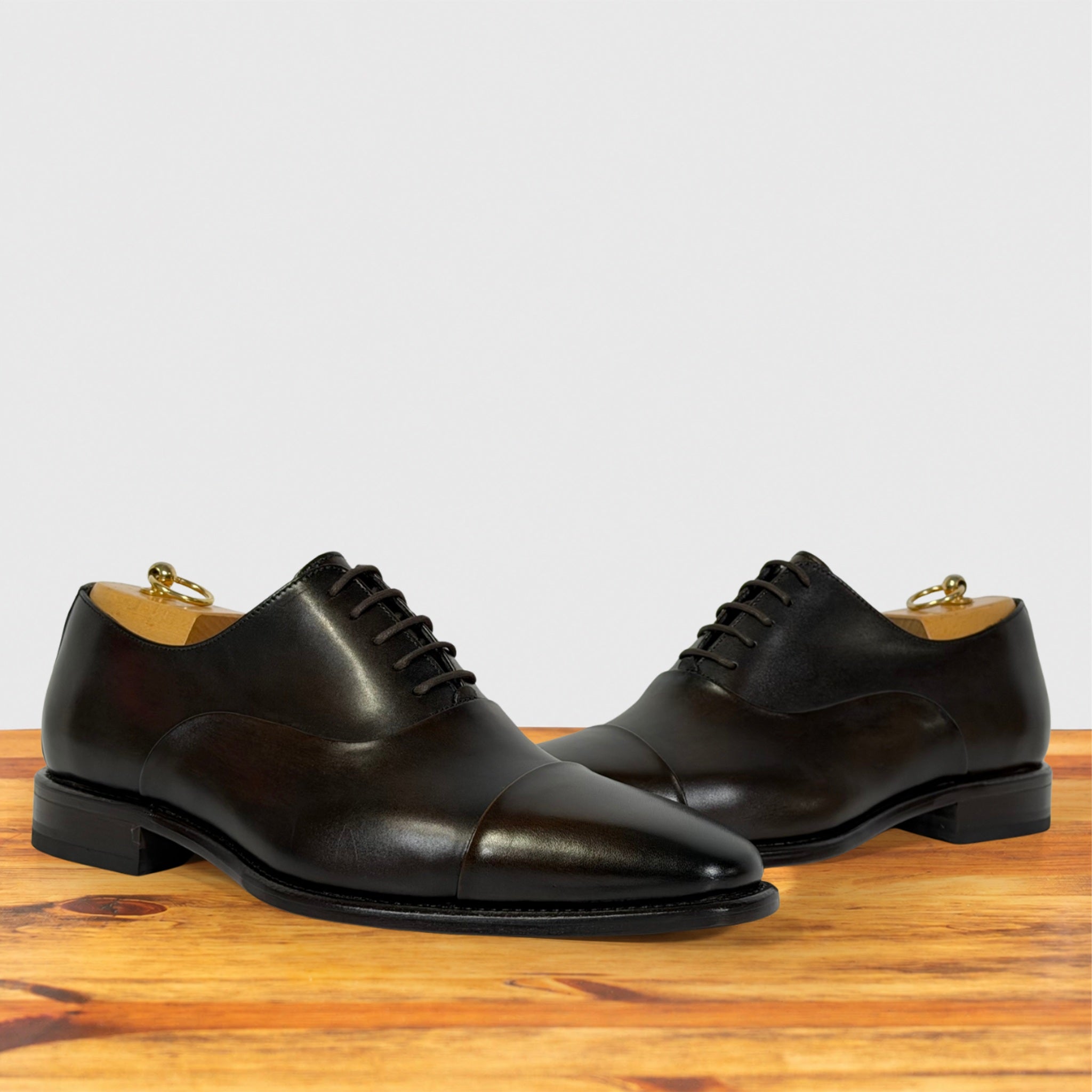 A pair of dark brown leather cap-toe oxford shoes with laces, displayed on a wooden surface against a light gray background.