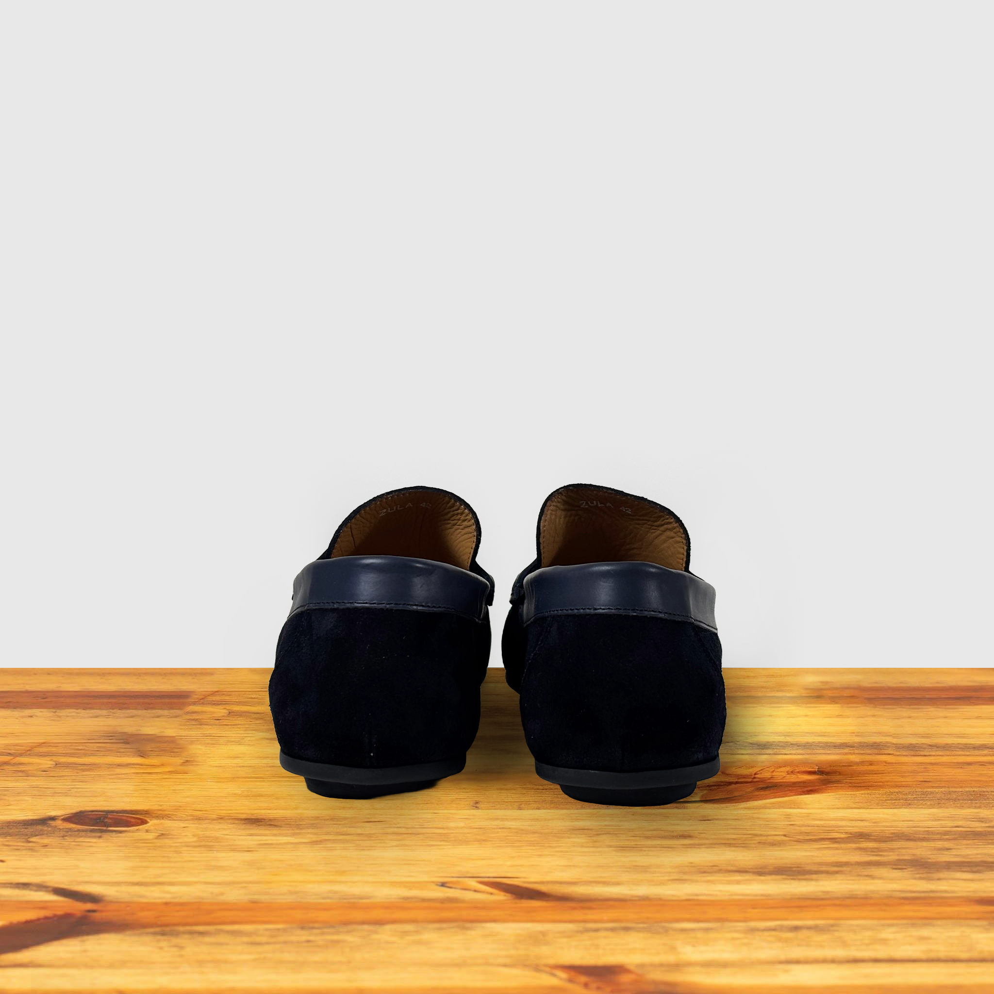 A navy blue suede penny loafer with a strap, displayed on a wooden surface.