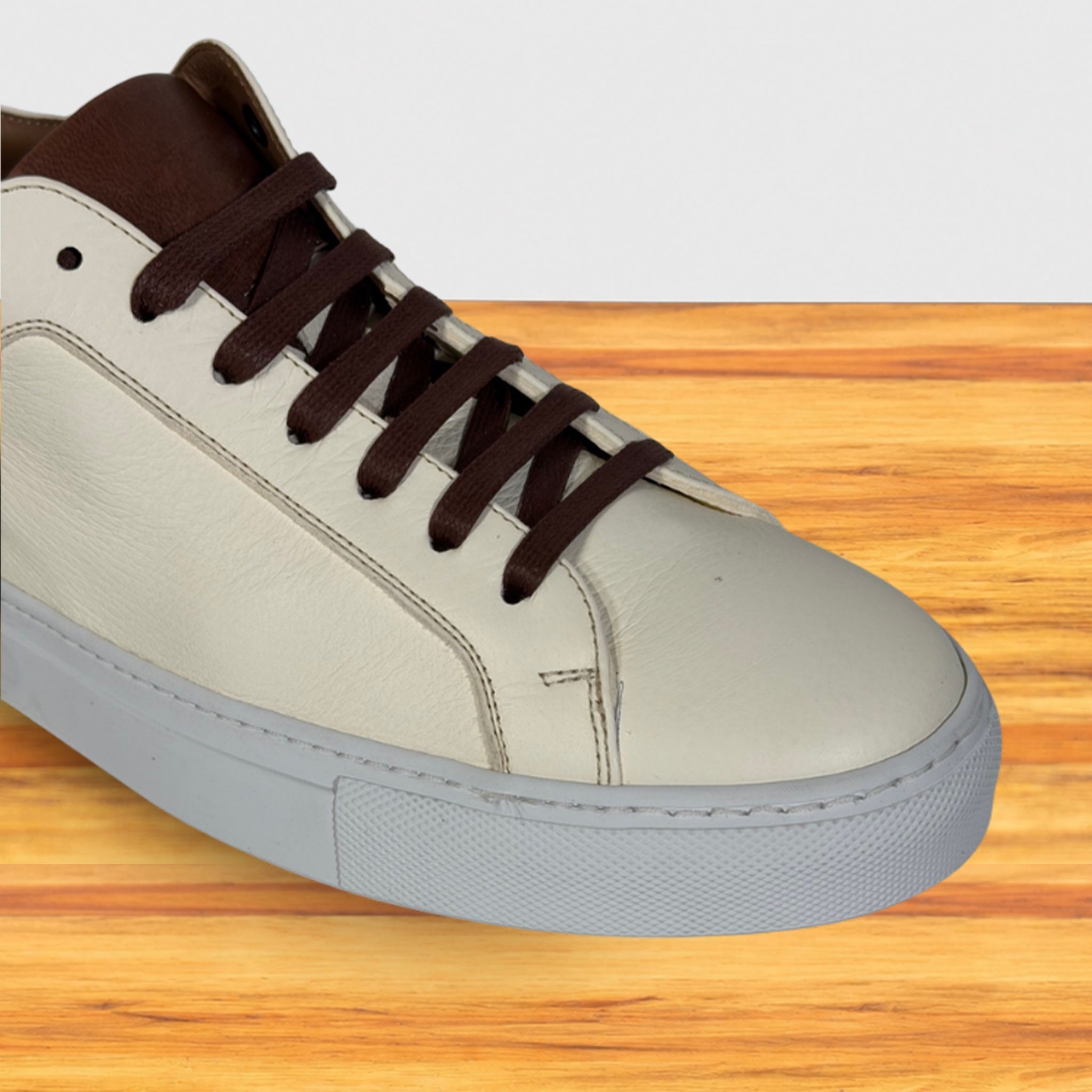 A low-top sneaker with a light cream leather upper, dark brown laces and collar, shown on a wooden surface against a white background.