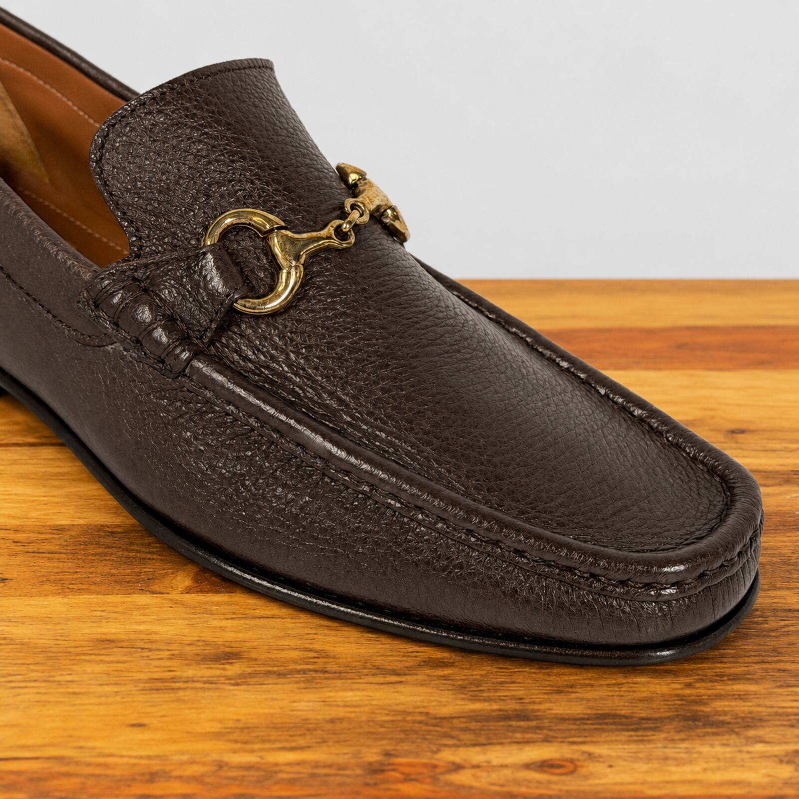 A close-up of a dark brown leather loafer with a gold buckle, resting on a wooden surface against a light gray background.