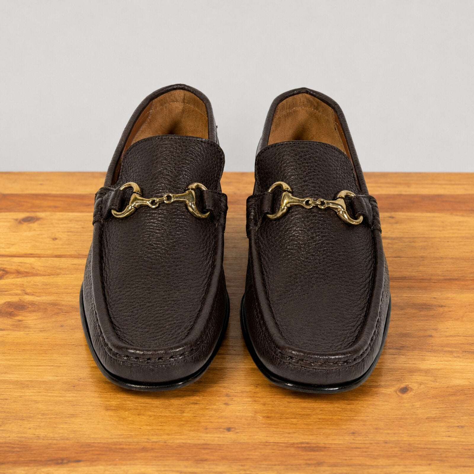 A pair of dark brown leather penny loafers with gold buckle on a wooden surface against a neutral light gray background.