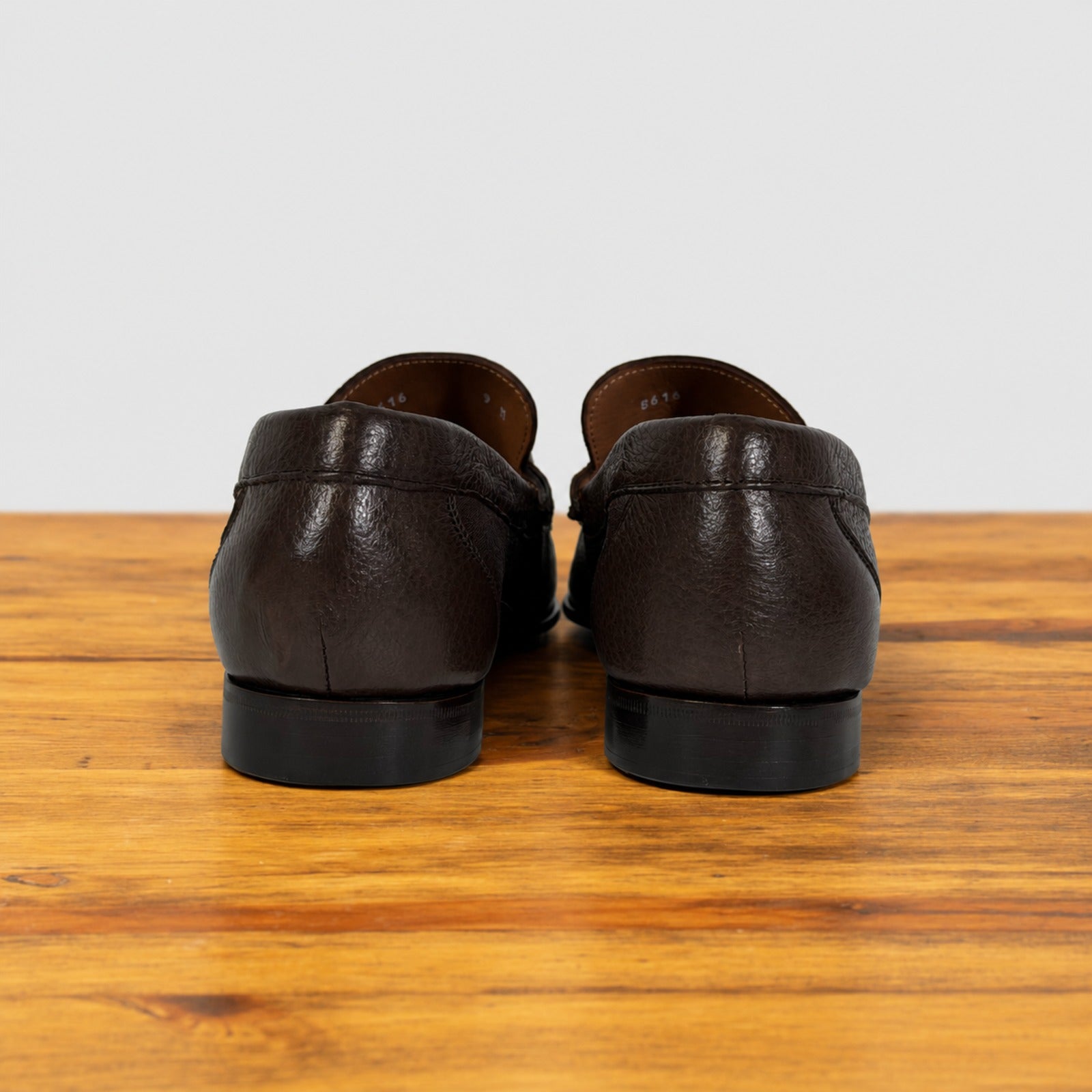 A pair of dark brown leather loafers shown from the back, resting on a wooden surface against a light gray background.