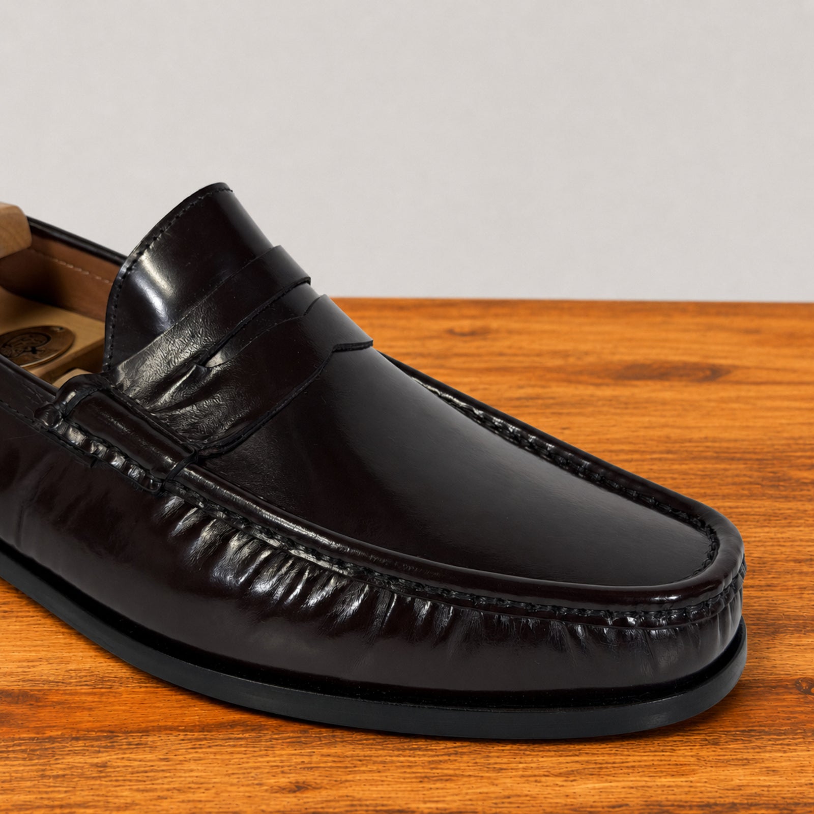 A close-up of a glossy burgundy leather penny loafer resting on a wooden surface against a light gray background.