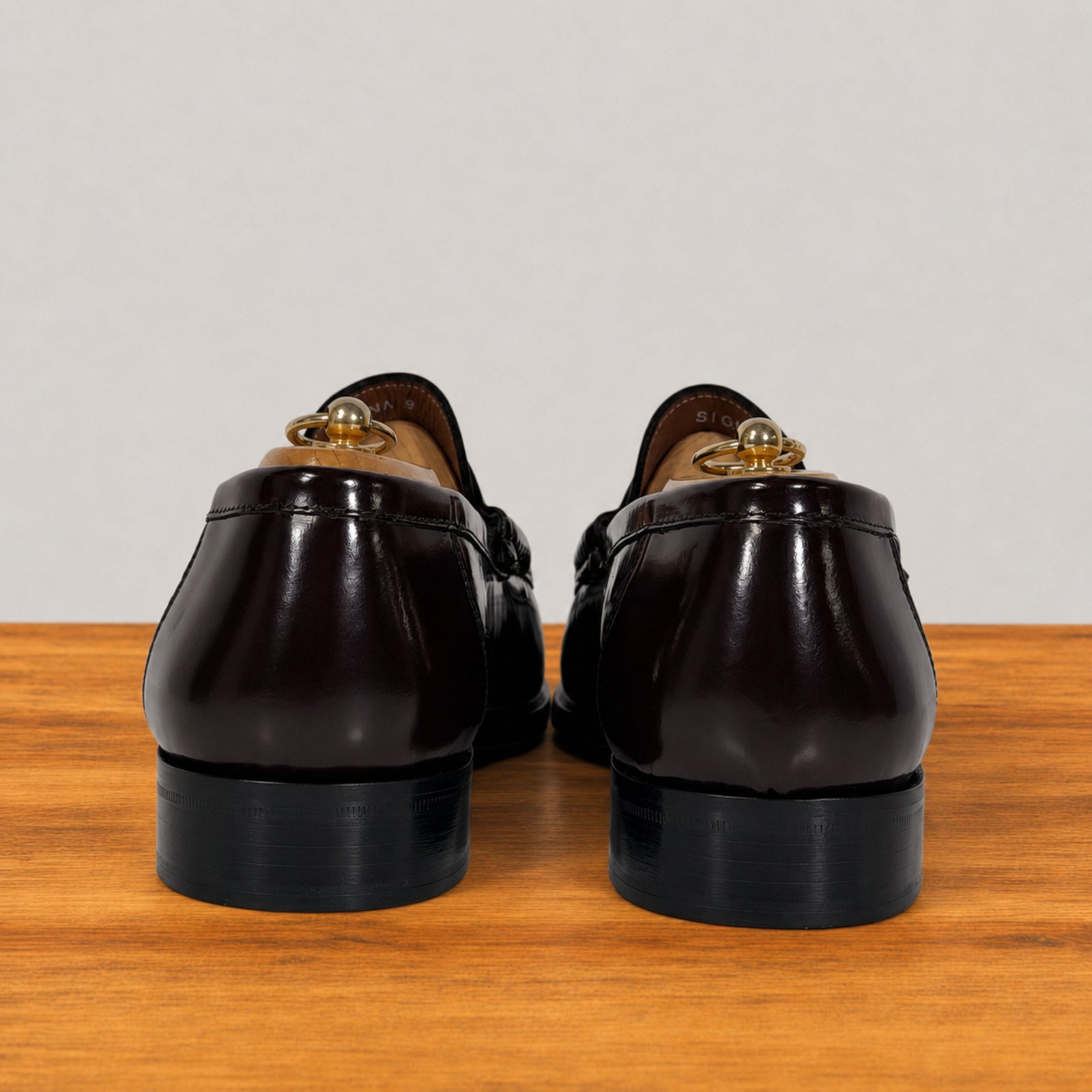 A pair of burgundy patent leather penny loafers, shown from the back on a wooden surface against a light gray background.
