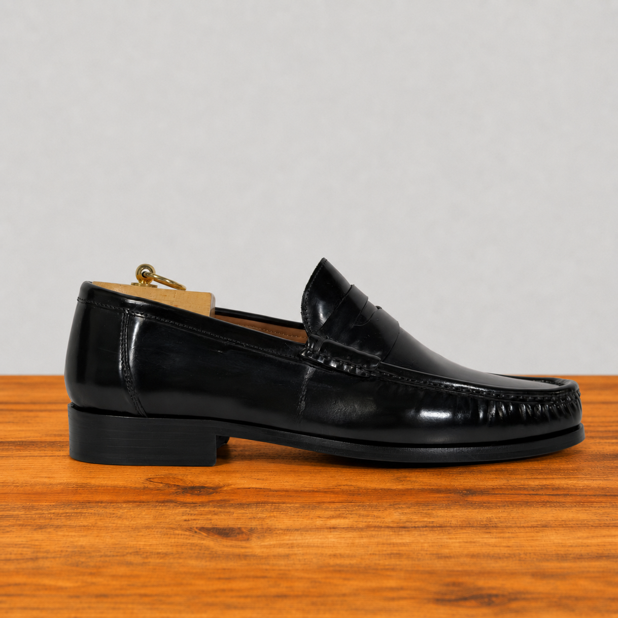 A single black leather penny loafer shoe with a low stacked heel, shown in profile on a wooden surface against a light gray background.
