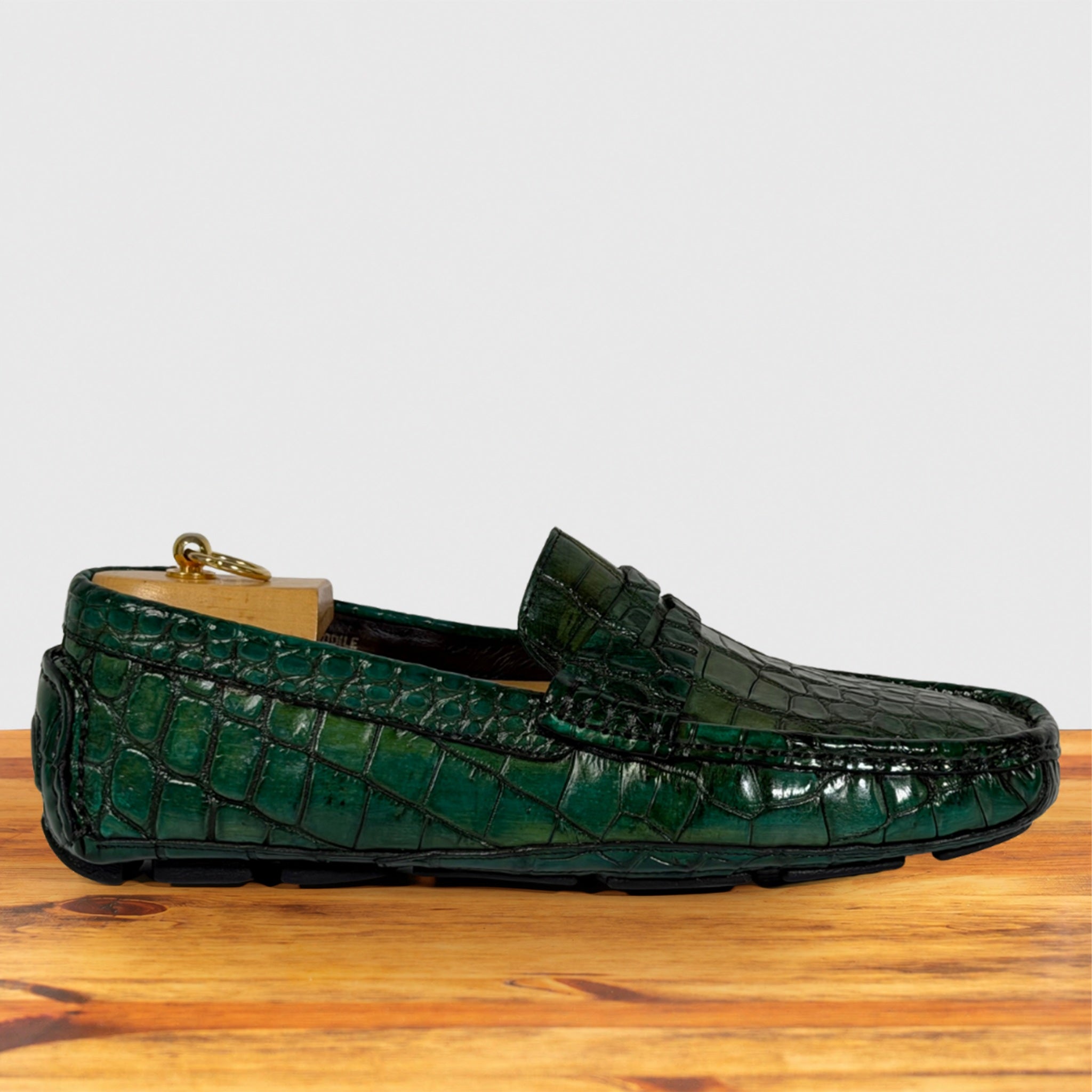 A single green crocodile-embossed loafer driving shoe resting on a wooden surface against a light gray background.