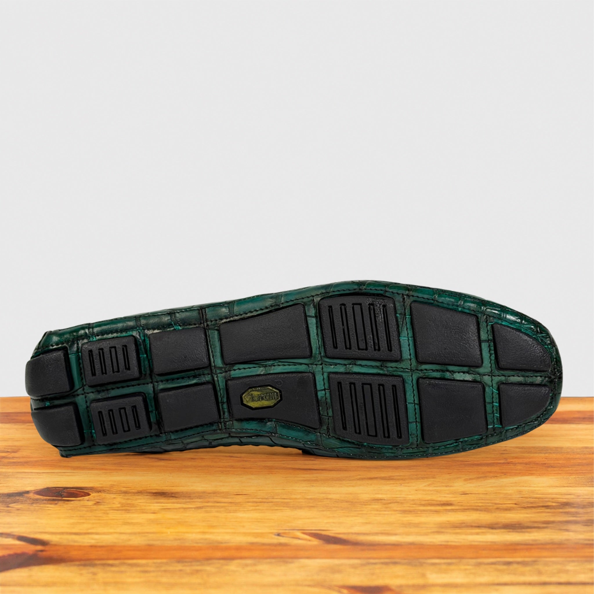 The outsole of a shoe with a green and black alligator-patterned tread, resting on a wooden surface against a light gray background.