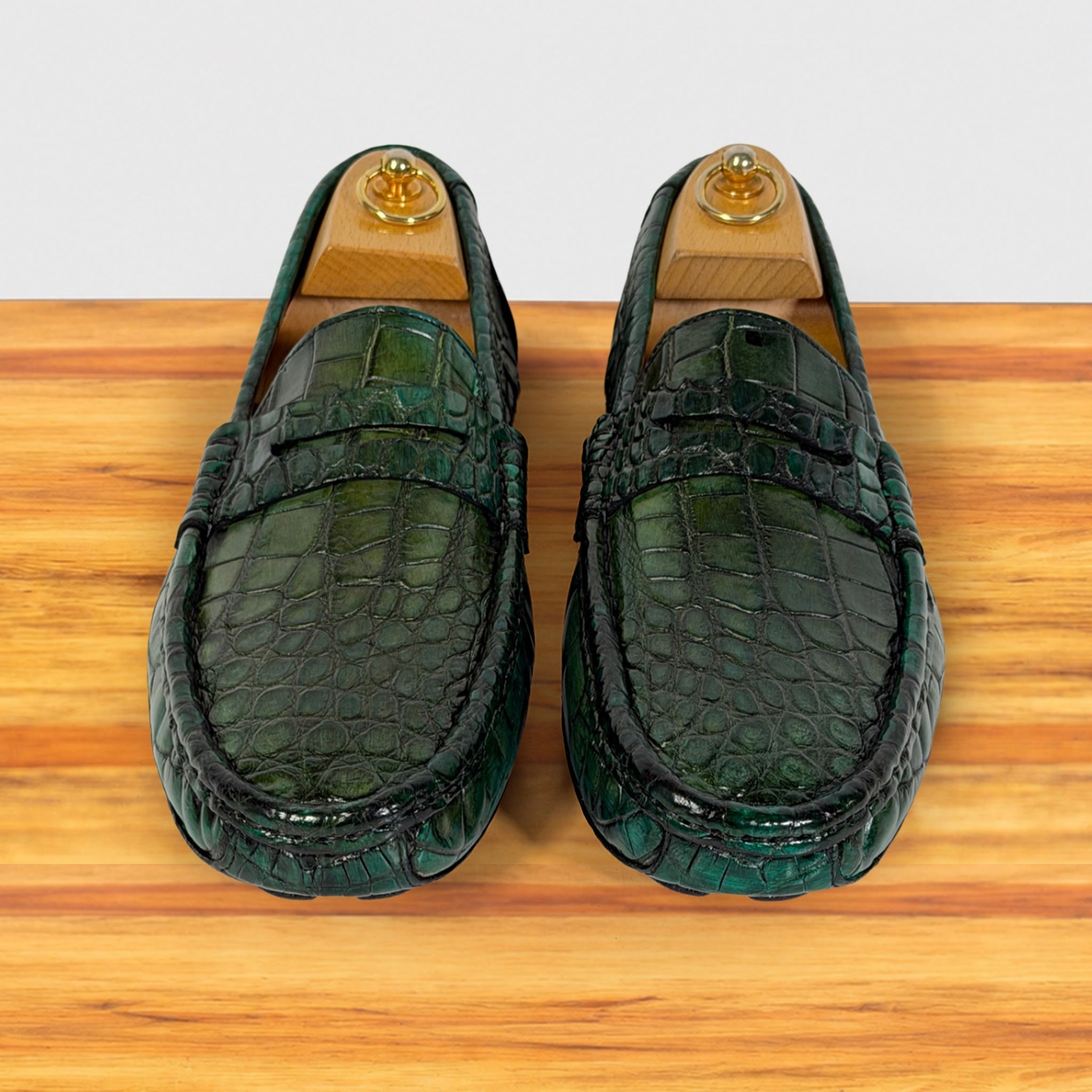 A pair of Emerald Green crocodile-embossed leather loafers displayed on wooden shoe stands over a light wood surface against a white background.