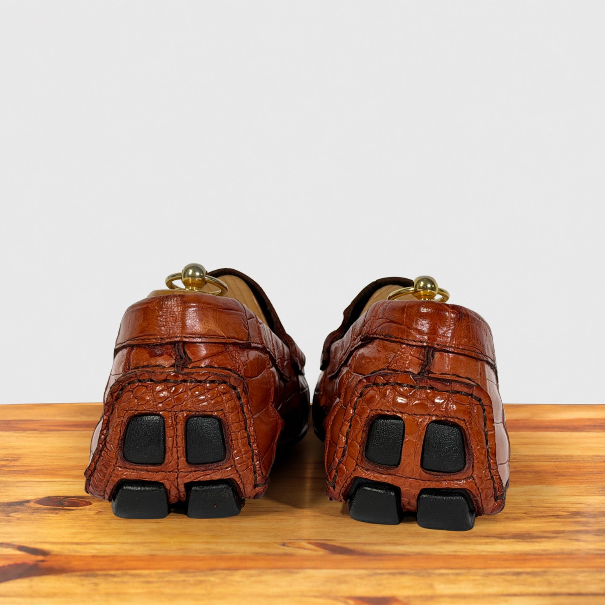 A pair of Burnt Orange crocodile-embossed leather loafers shown from the back, resting on a wooden surface against a light gray background.