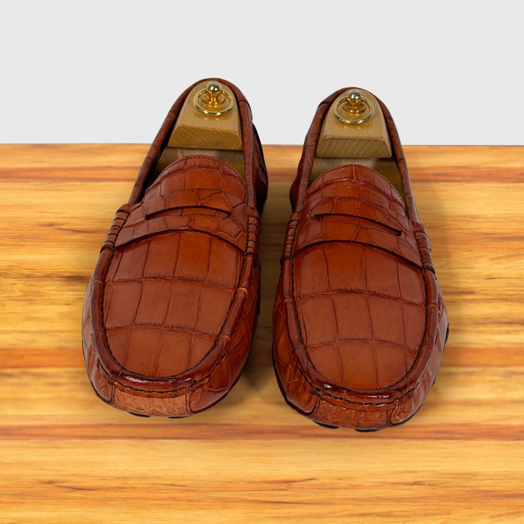 A pair of Burnt Orange crocodile-embossed leather penny loafers resting on a light wood surface against a white background.