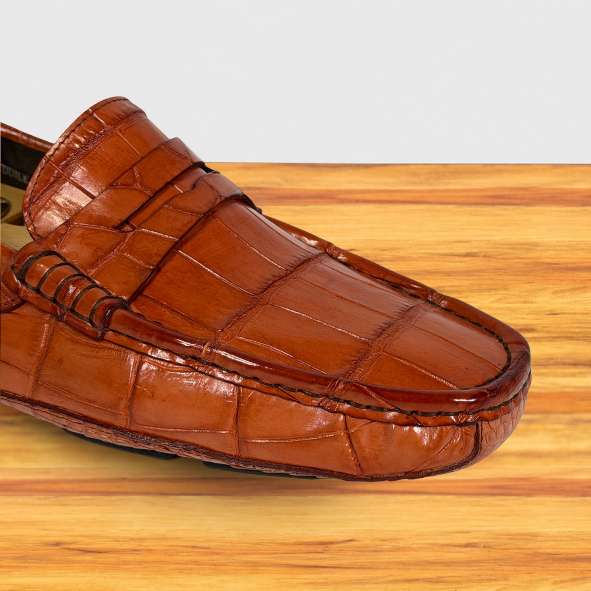 A single Burnt Orange crocodile-embossed leather loafer shown in a close-up studio-style shot on a light wood surface against a pale gray background.
