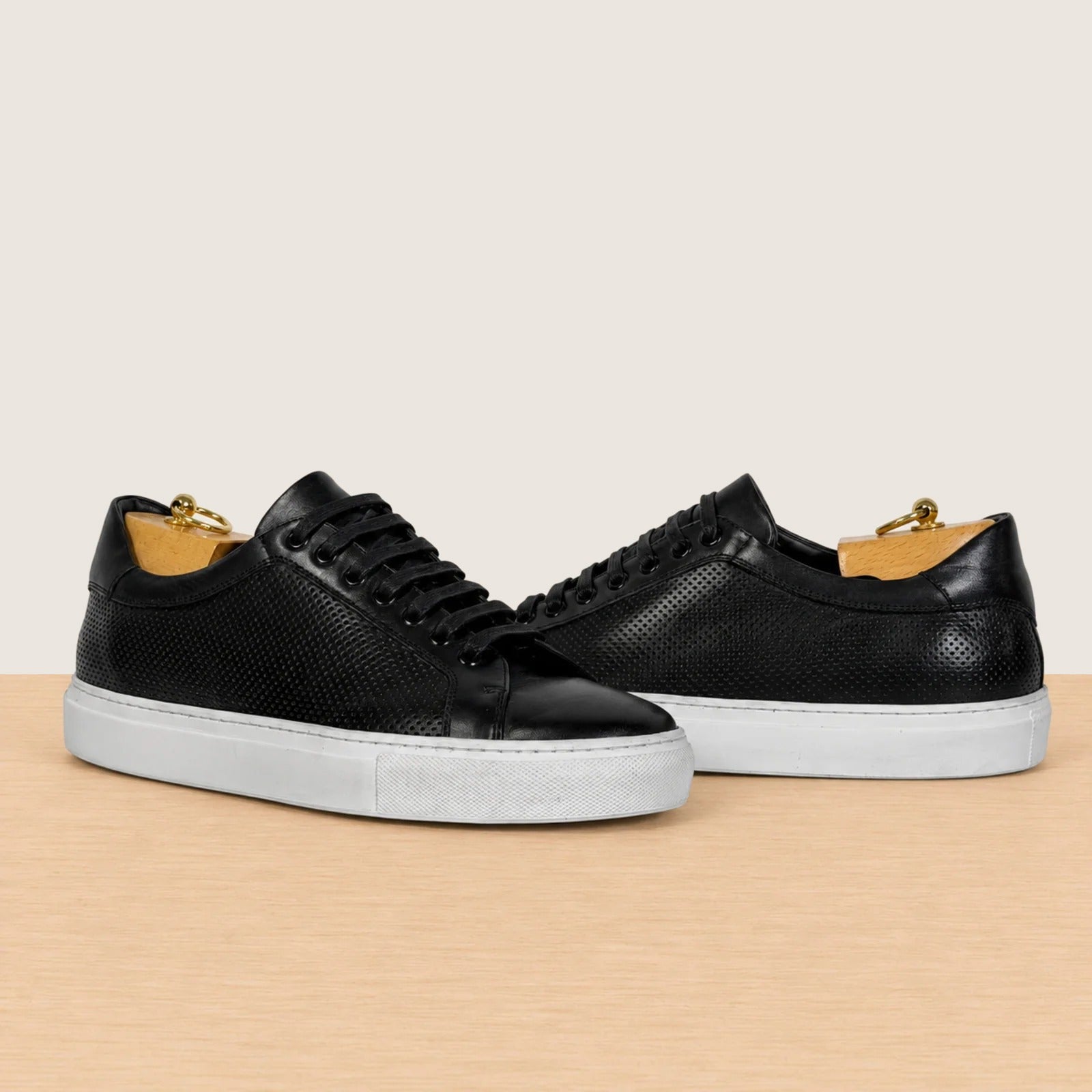 A pair of black leather low-top sneakers with textured perforated uppers, black laces, and thick white platform soles, displayed on a light wood surface against a warm beige background.