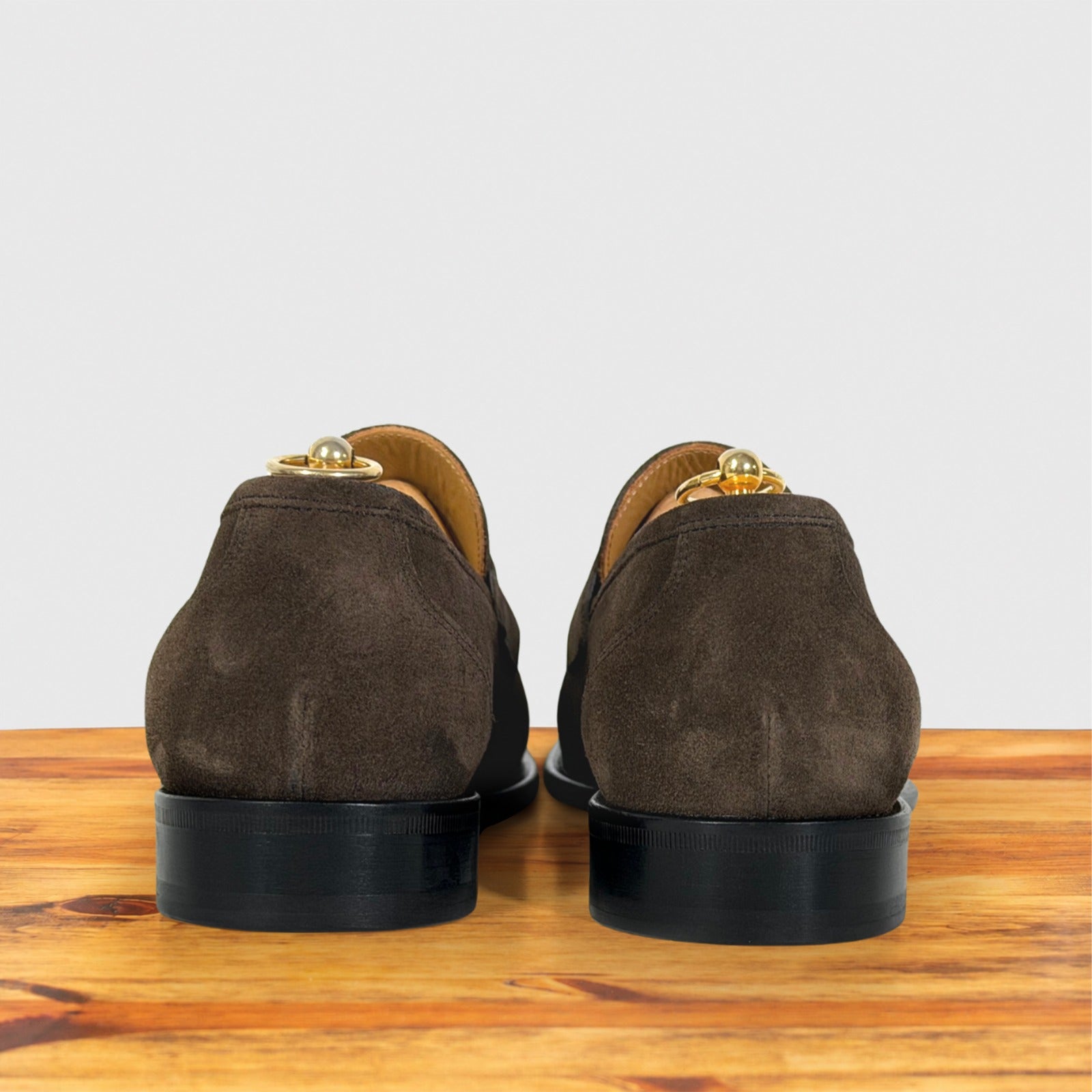 A pair of dusty brown suede loafers and black soles, shown from the back on a wooden surface against a light gray background.
