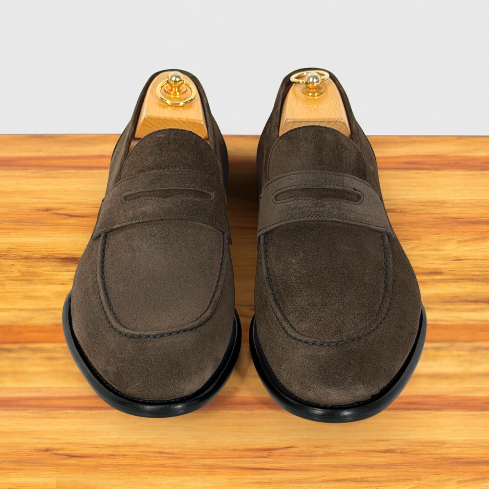 A pair of dusty brown suede penny loafers with black soles, displayed on wooden shoe trees on a wooden surface against a light gray background.