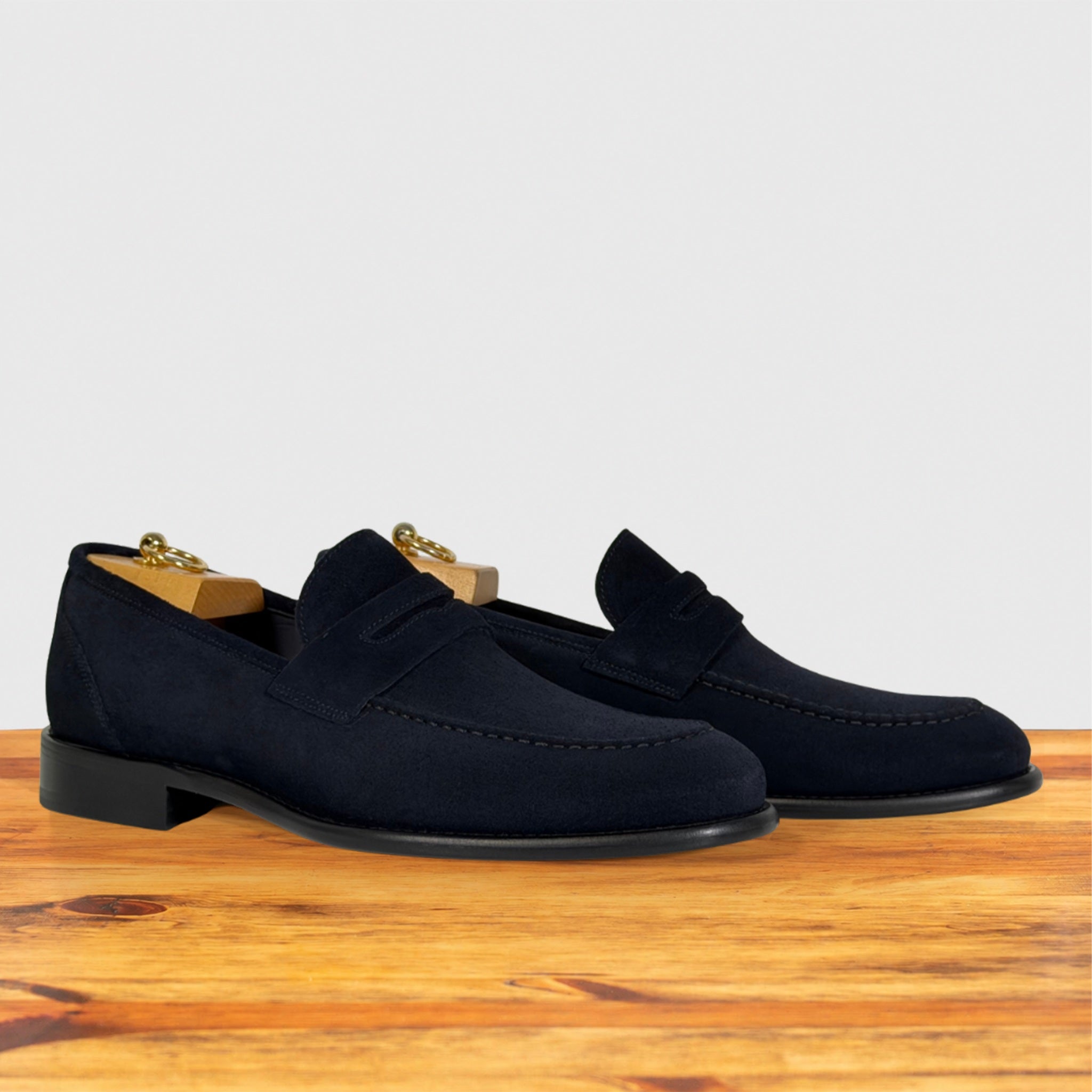 A pair of navy blue suede penny loafers resting on a wooden surface against a light gray background.