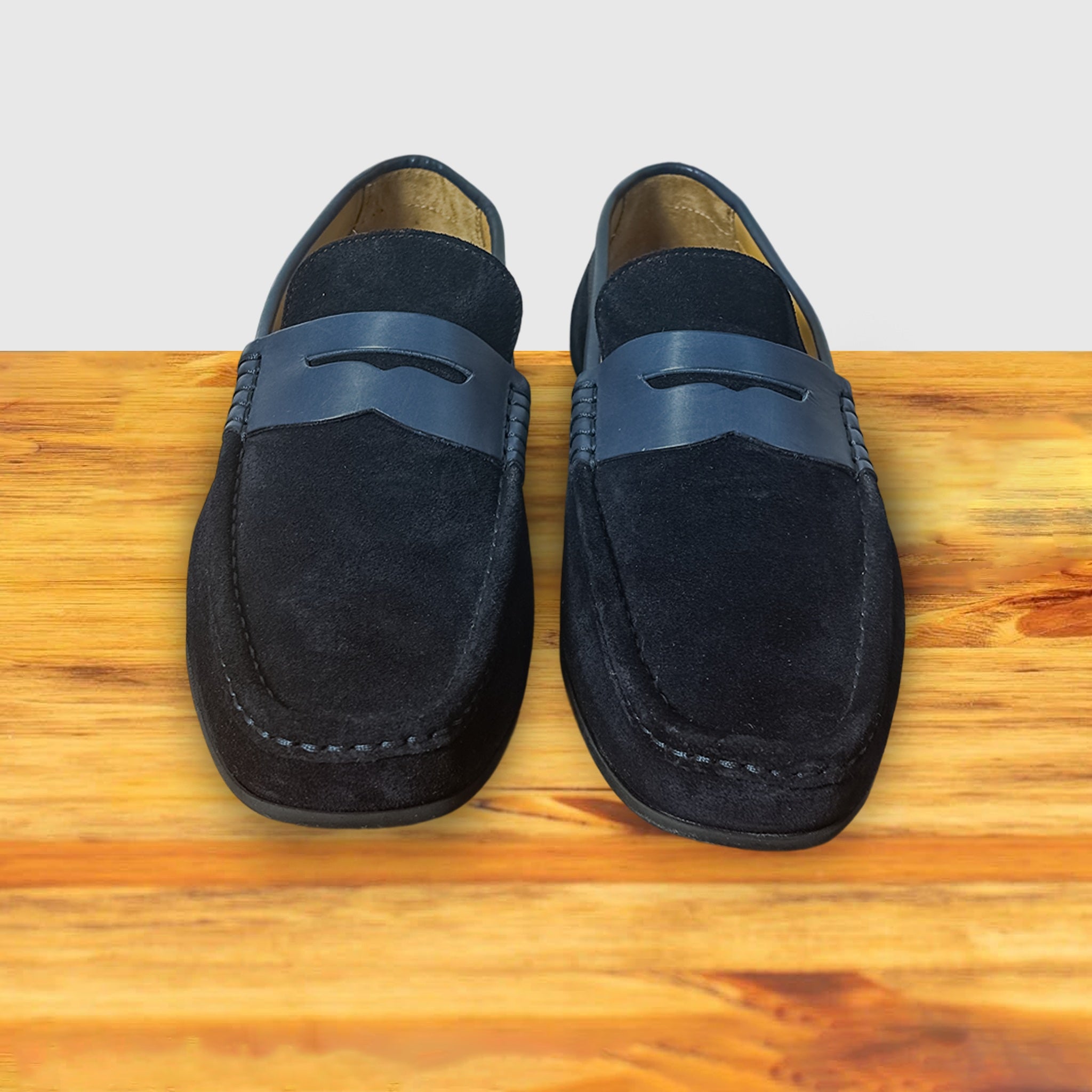 A navy blue suede penny loafer with a strap, displayed on a wooden surface.