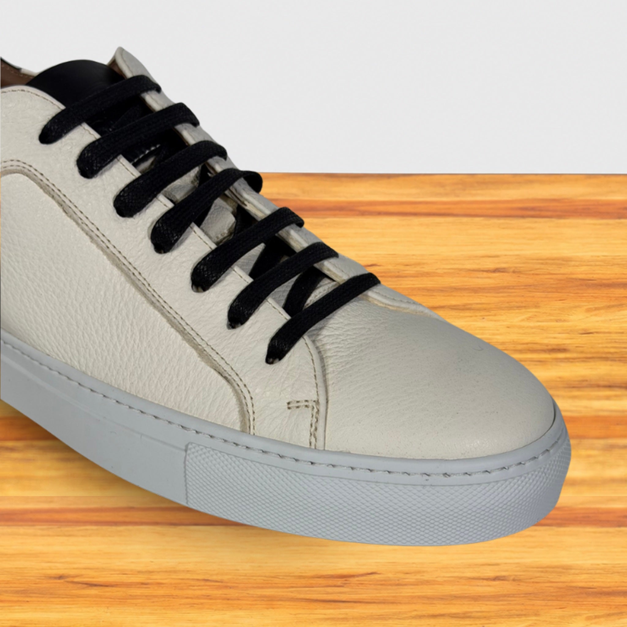 A lwhite ow-top sneaker with black laces, and a light gray sole, shown on a wooden surface against a white background.