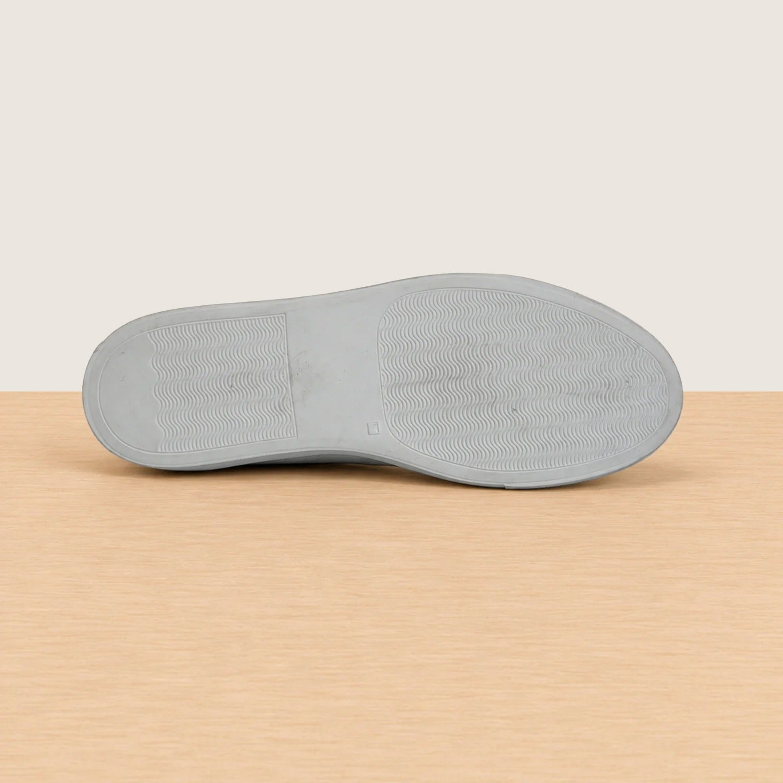 The outsole of a slip-on shoe, showing textured tread patterns, resting on a light wood surface against a beige background.