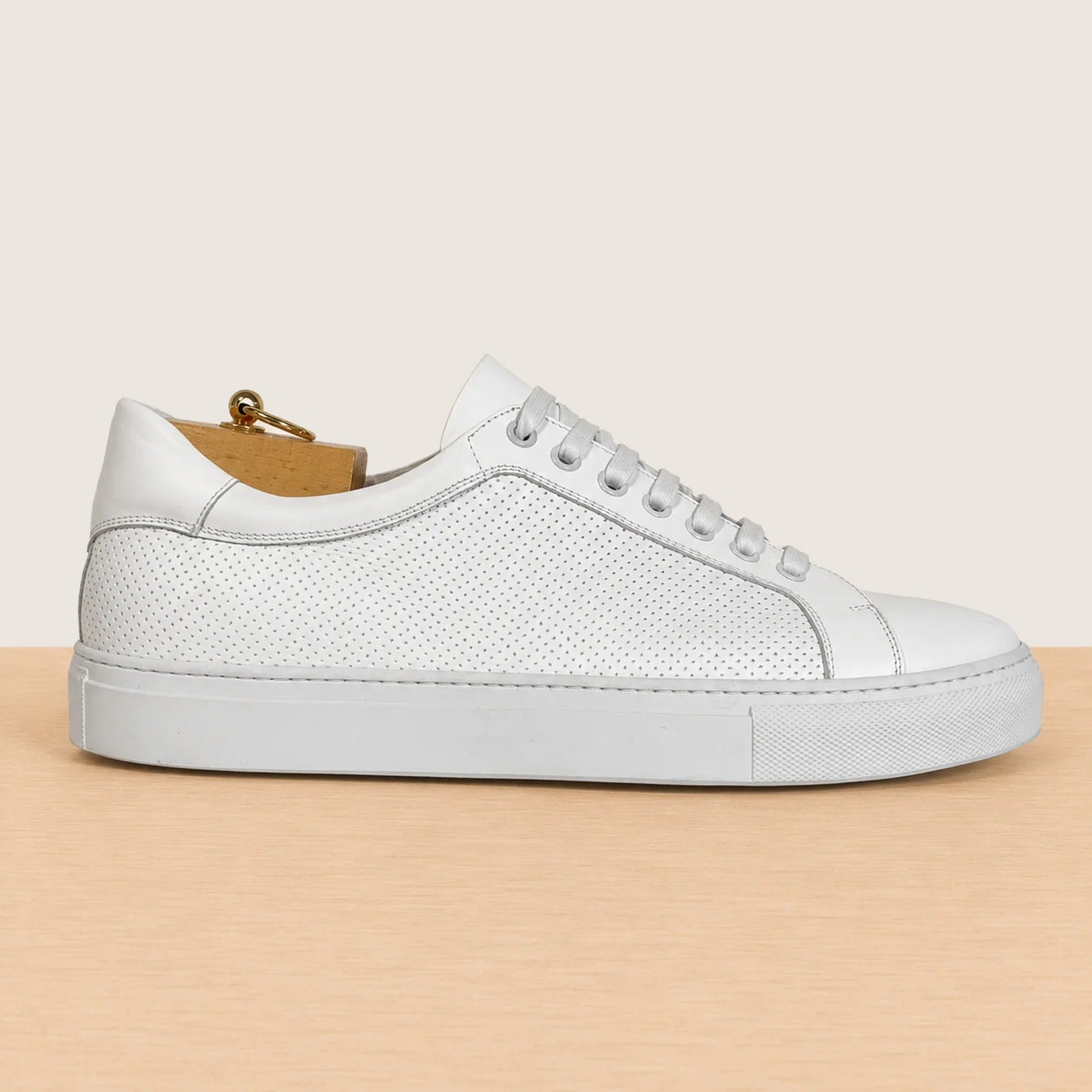 A white leather low-top sneaker with perforated detailing, white laces, and a thick white sole, shown in profile on a light wood surface against a warm beige background.