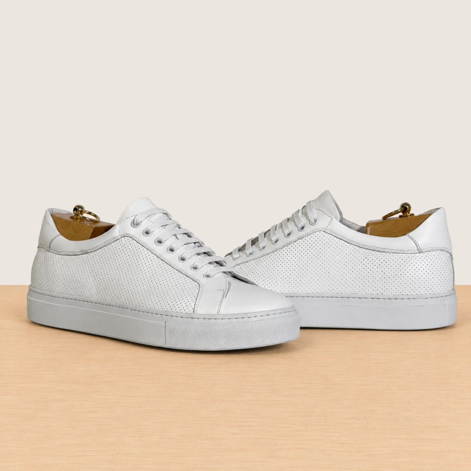 A pair of white leather low-top sneakers with perforated detailing, shown on a light wood surface against a warm beige background.