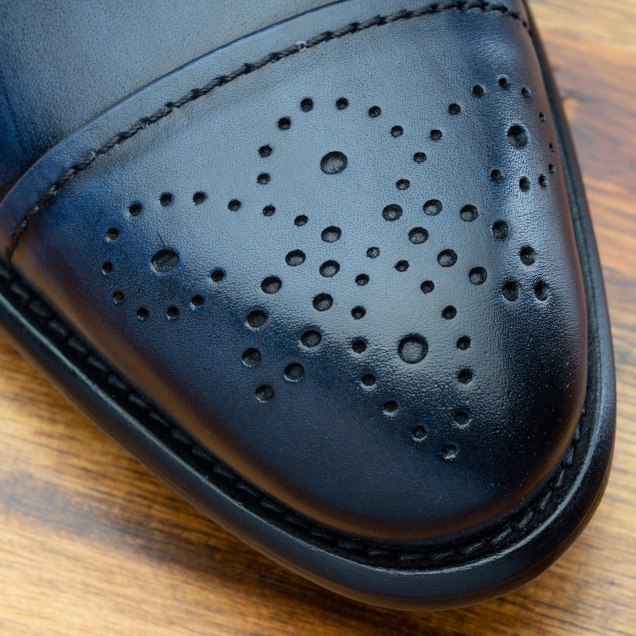Close-up of a dark blue leather dress shoe toe featuring decorative perforations and stitching, resting on a wooden surface.