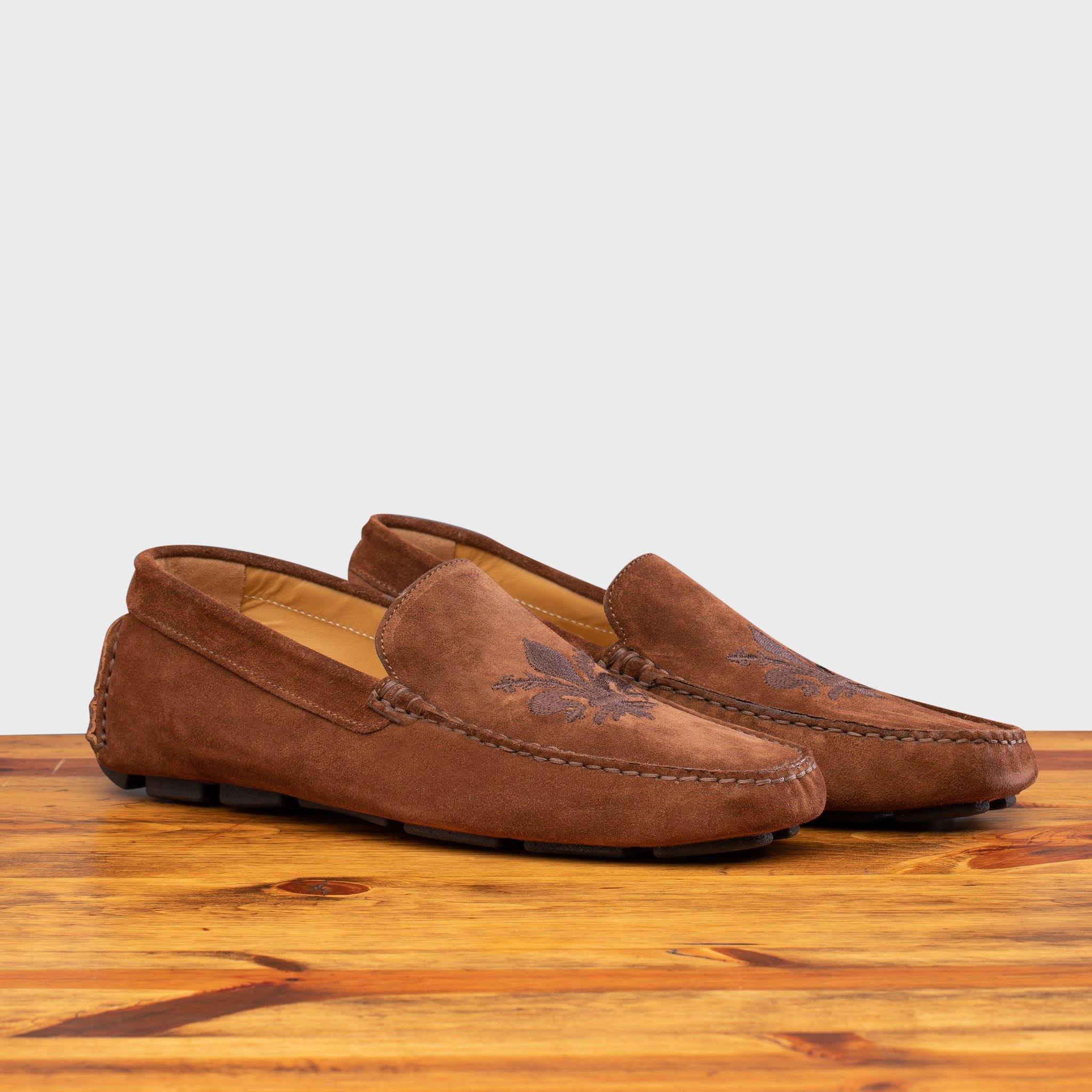Pair of 5303 Calzoleria Toscana Brick Venetian Suede Driver with brown embroidered fleur di Lis on top of a wooden table