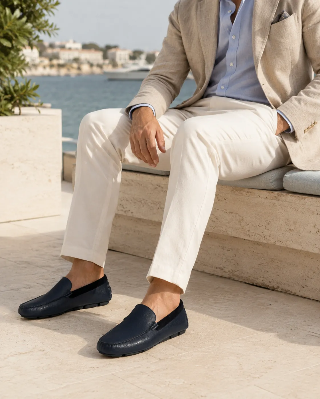 Man in a linen blazer, light blue shirt and cream trousers seated on a travertine bench above the Mediterranean, wearing handcrafted navy Calzoleria Toscana Gioi saffiano leather drivers.