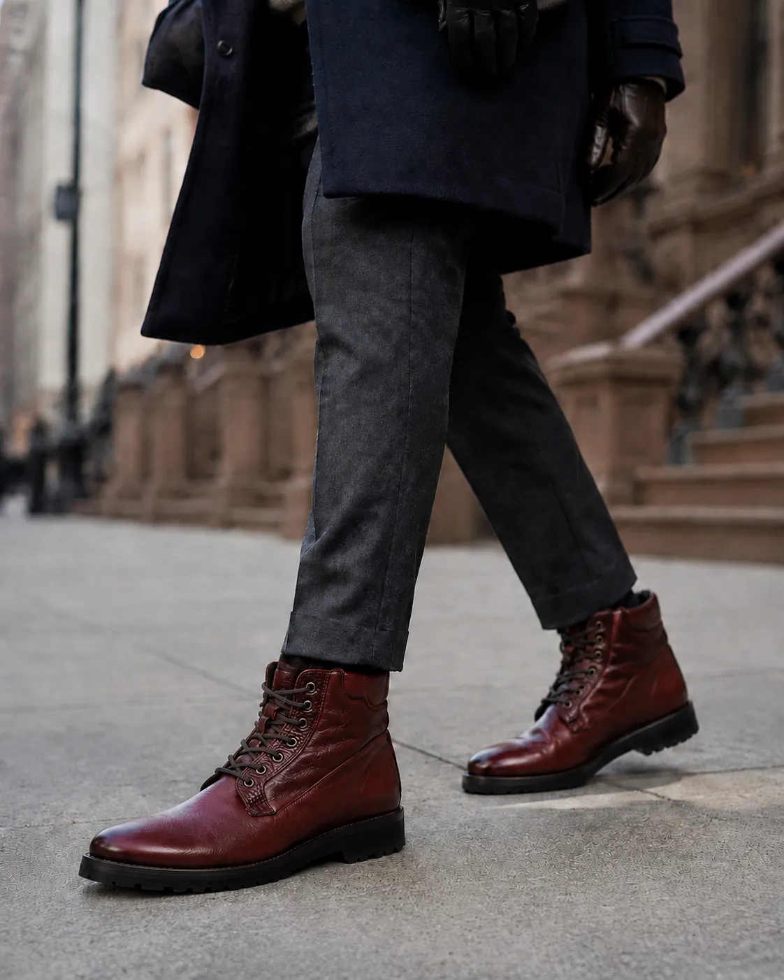 Man in a navy overcoat, charcoal trousers and leather gloves walking a city sidewalk, wearing handcrafted burgundy Calzoleria Toscana 3236 shearling-lined lace-up boots.
