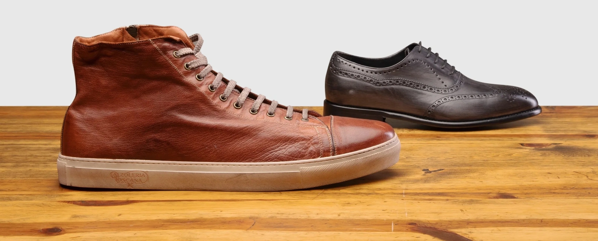 Two Calzoleria Toscana men's shoes in US size 14–18 displayed on a wooden surface: a cognac brown high-top leather sneaker with a cream rubber sole on the left, and a grey hand-burnished wingtip oxford with brogue detailing on the right.