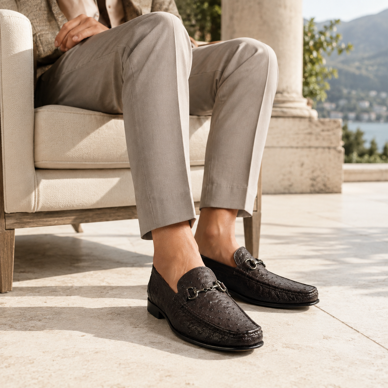 Man in light stone trousers seated on a sunlit lakeside terrace, wearing handcrafted dark brown Calzoleria Toscana 3238-M ostrich dress loafers with polished nickel bits.