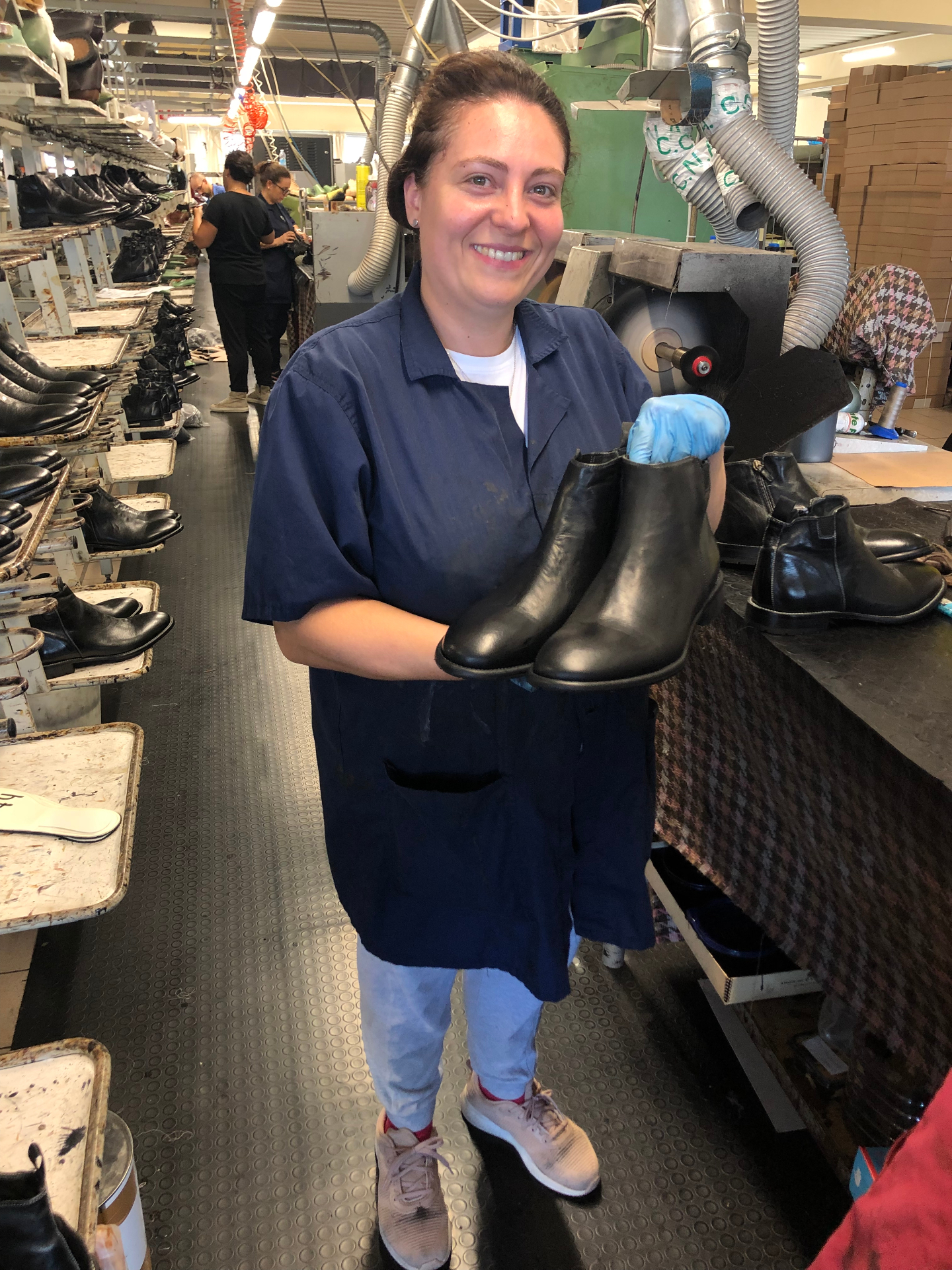 A Toscana artisan on the finishing line smiling as she holds up a pair of just-finished black leather Chelsea boots