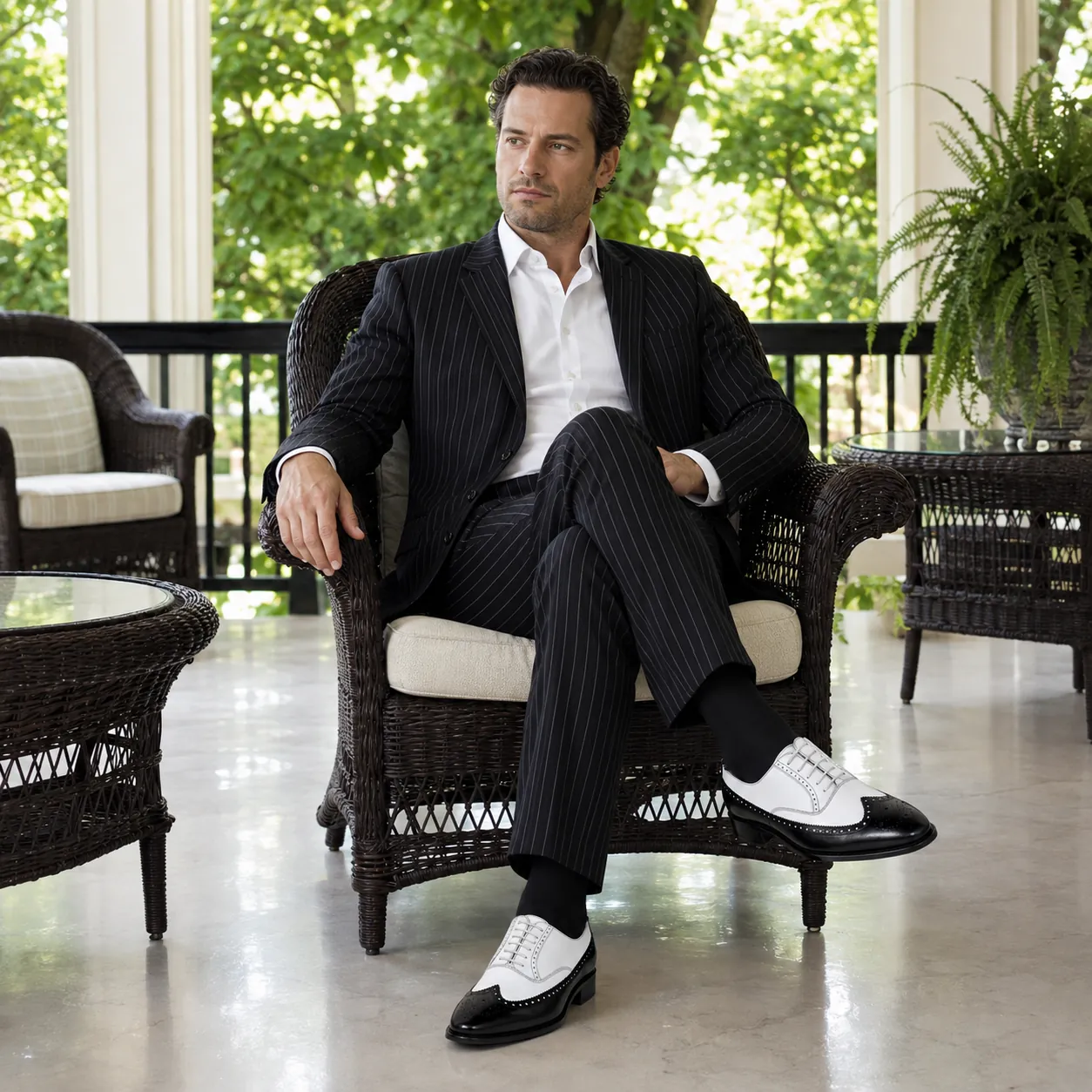 Man in a black pinstripe suit and white shirt seated in a wicker armchair on a sunlit veranda, wearing handcrafted black and white Calzoleria Toscana Caldes spectator wingtip oxfords.