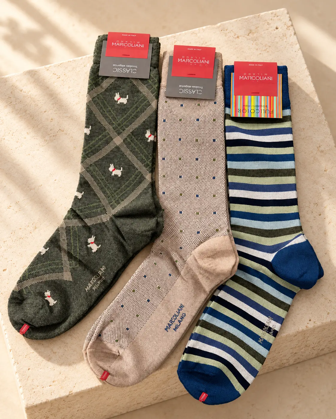 Three Marcoliani Italian dress socks laid on limestone: green argyle with terriers, beige with small dots, and blue and green stripes.