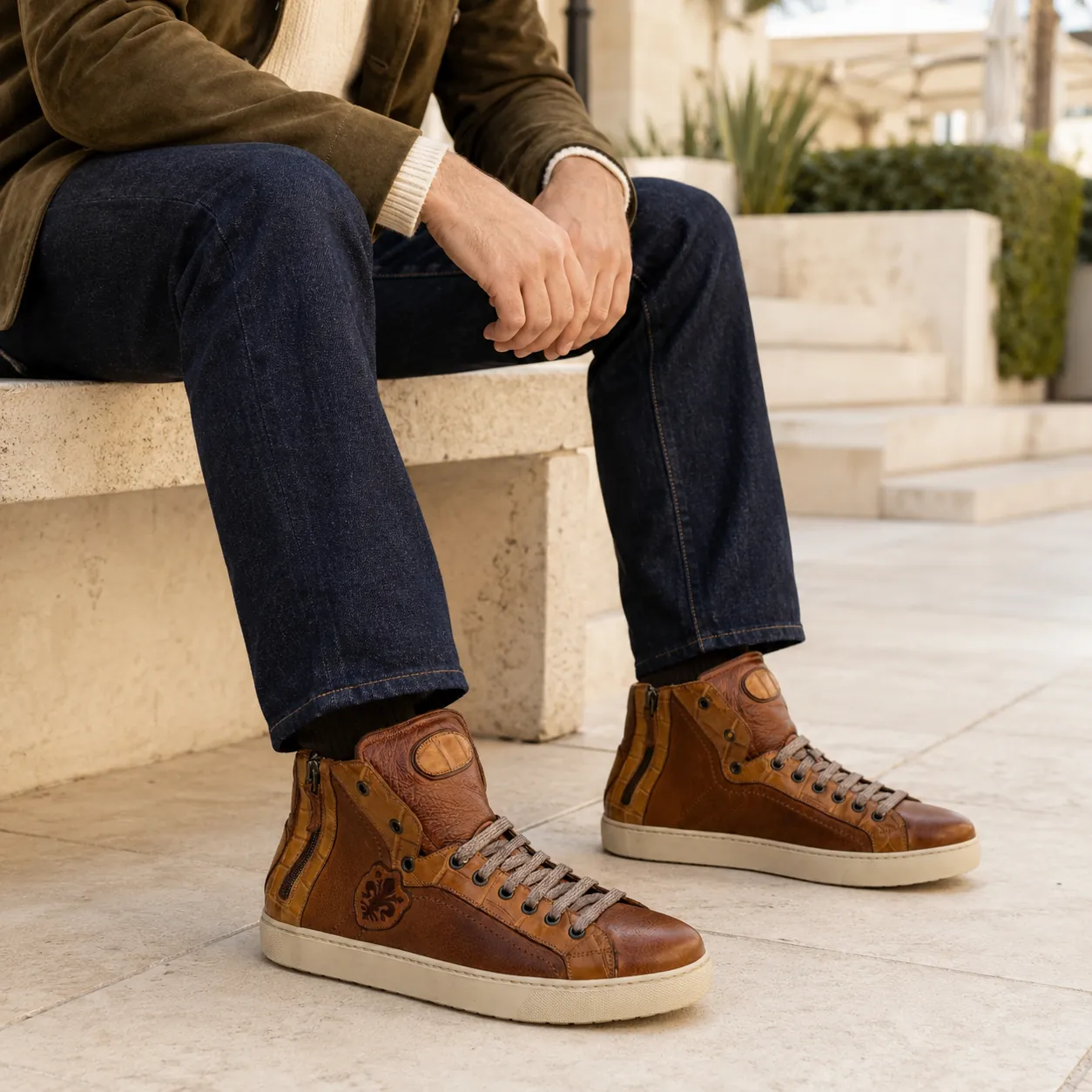 Man in an olive suede jacket, cream knit and indigo jeans seated on a limestone bench, wearing handcrafted cognac Calzoleria Toscana Goni dip-dyed crocodile high-top sneakers.