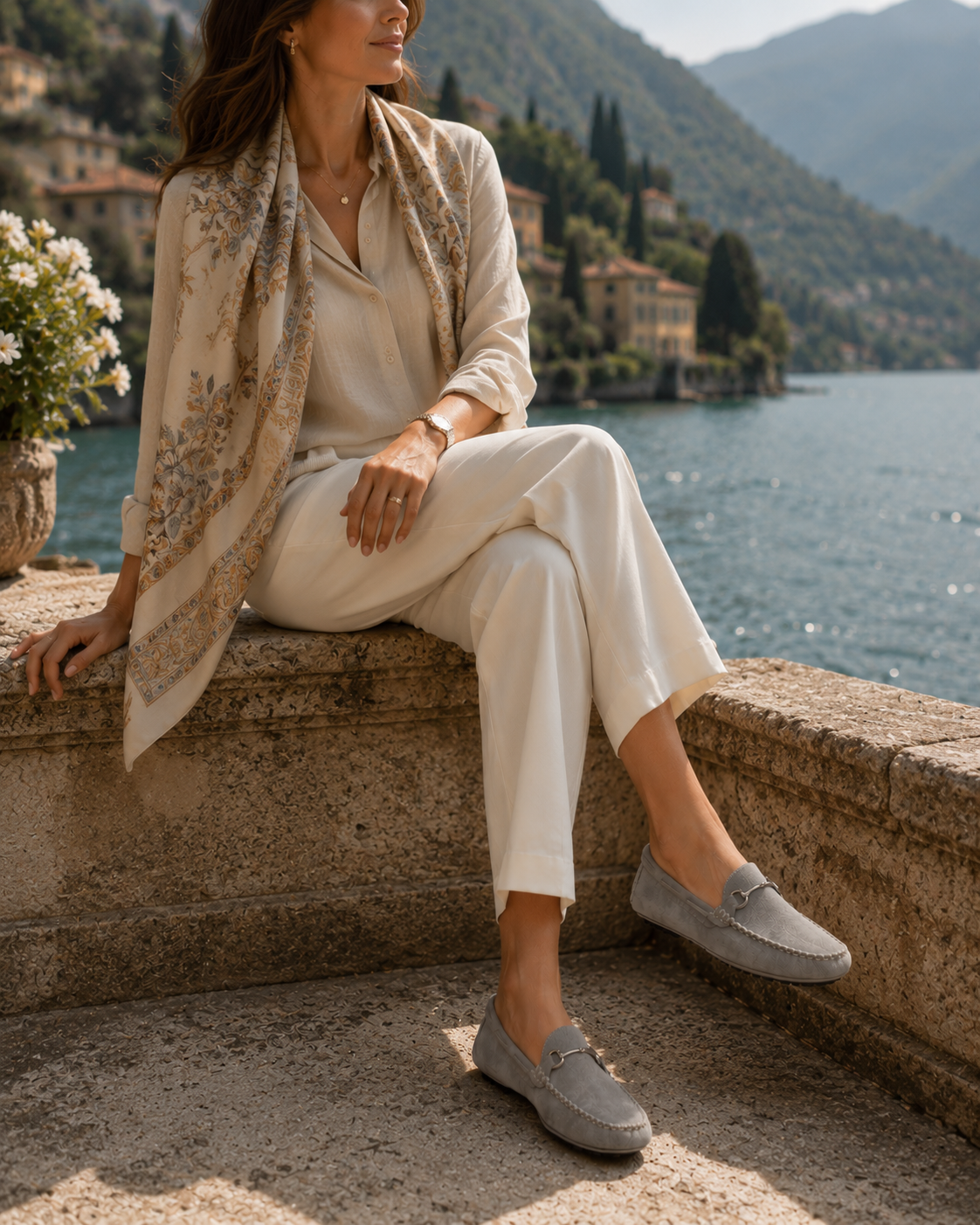 A woman in ivory linen wide-leg trousers and a soft camel cashmere sweater seated on warm travertine steps in a sunlit Tuscan piazza at golden hour, wearing handcrafted velour suede Italian driving moccasins by Calzoleria Toscana.