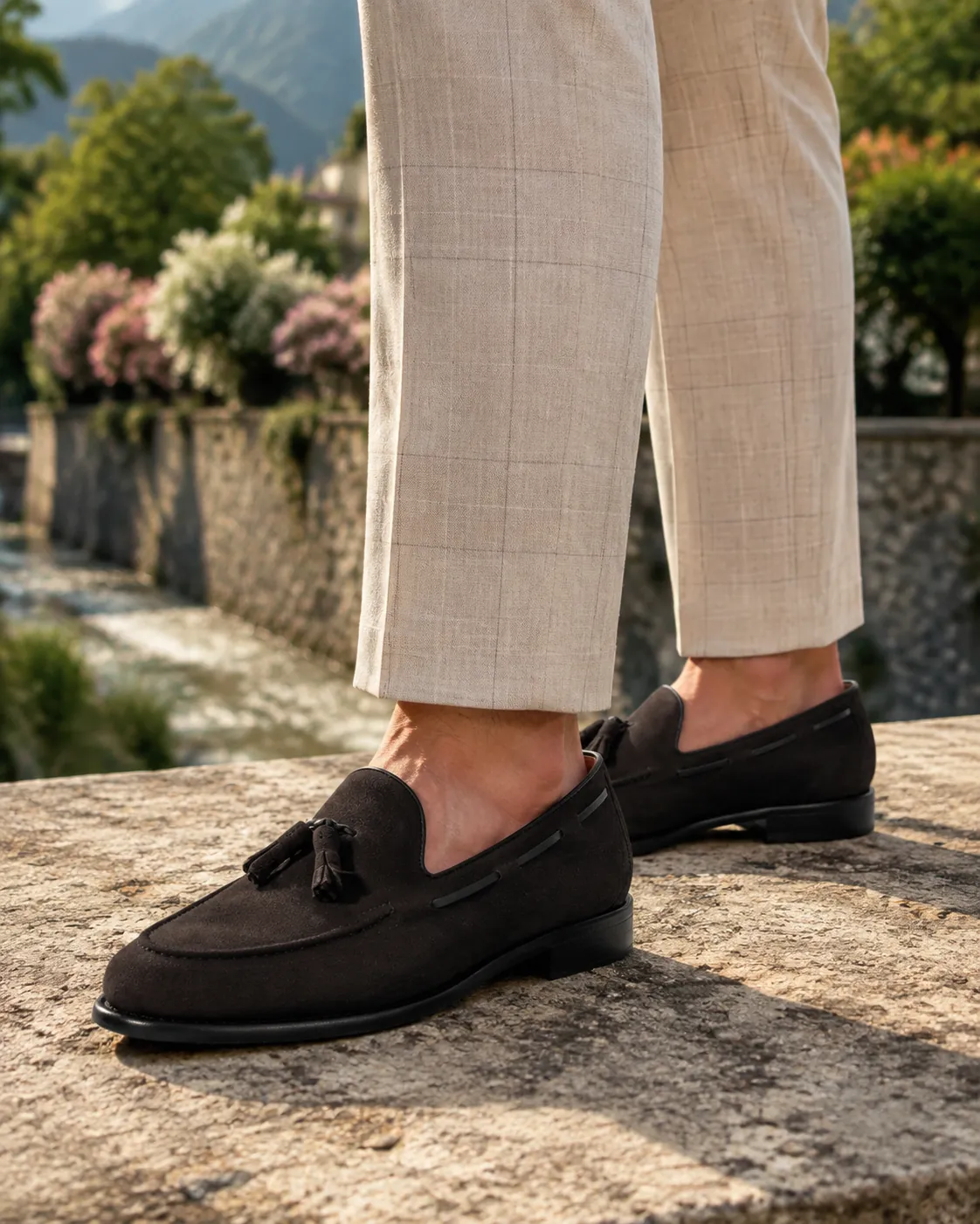 Man in cream windowpane linen trousers standing on a sunlit stone wall above an Italian lakeside garden, wearing handcrafted dark brown Calzoleria Toscana Bormio-S velour suede tassel loafers.