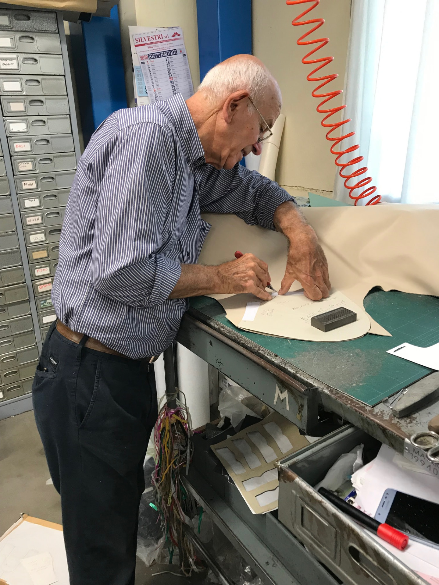 A master artisan at his workbench marking a leather panel by hand beside a steel cutting die in the Tuscan workshop