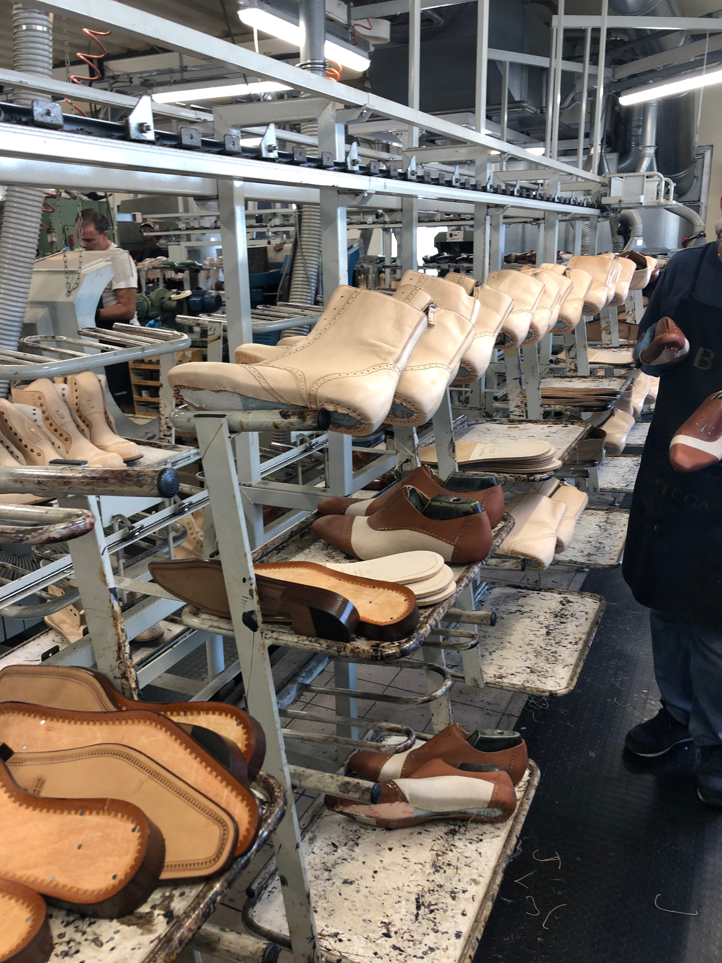 Rows of cream and tan leather uppers on wooden lasts lined up along the workshop racks, with artisans working at stations down the Tuscan factory floor