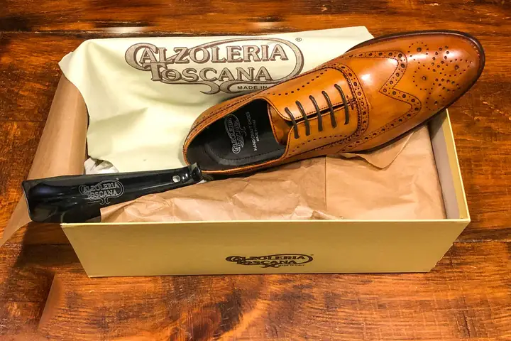 A cognac brown Calzoleria Toscana wingtip oxford with a shoehorn resting in its signature gold Made in Italy shoe box