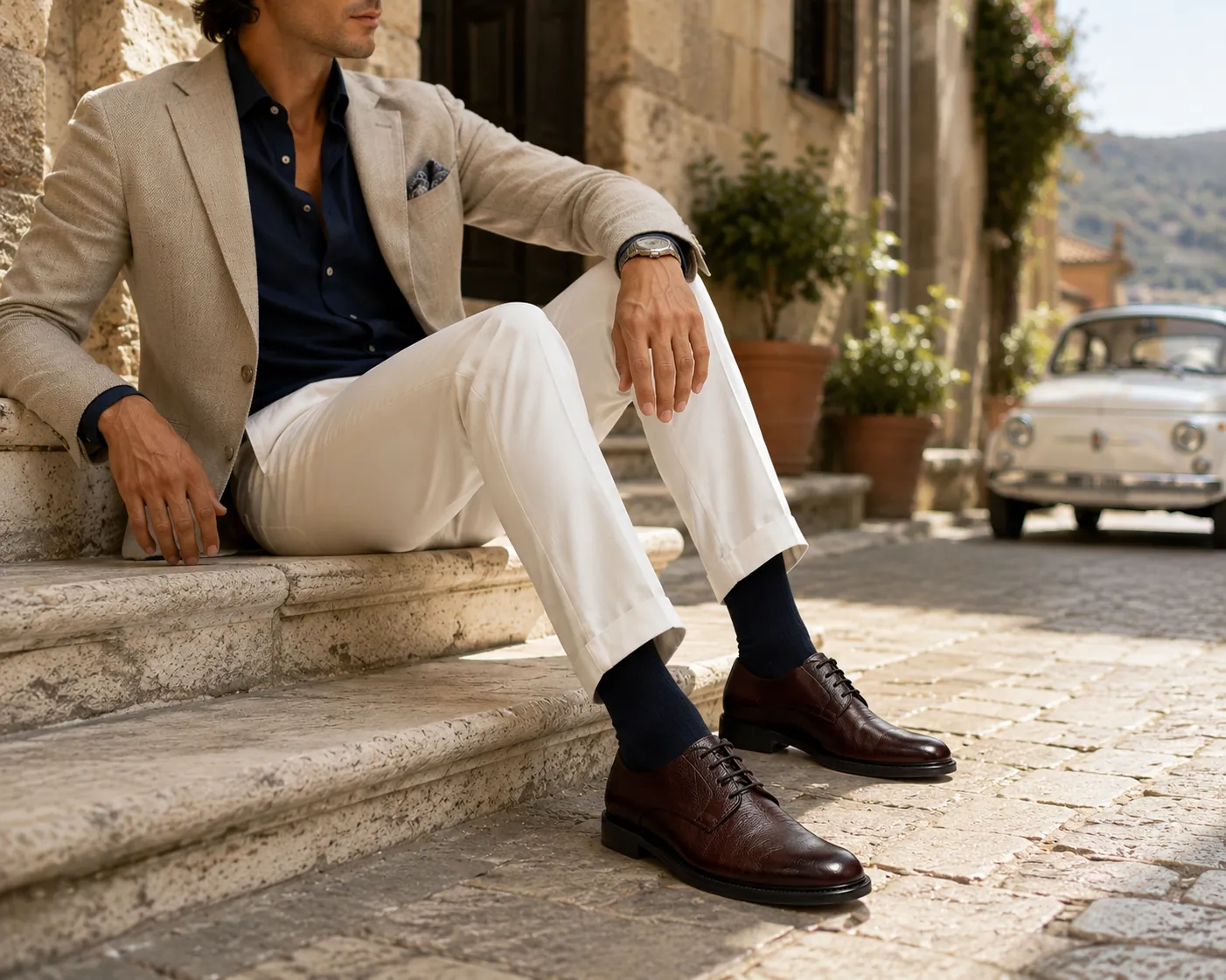 Man in a linen blazer, navy shirt and cream trousers seated on stone steps in an Italian village, wearing handcrafted brown Midas leather derbies by Calzoleria Toscana.