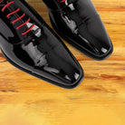 A close-up of a pair of glossy black patent leather dress shoes with bright red laces, resting on a warm wooden surface.