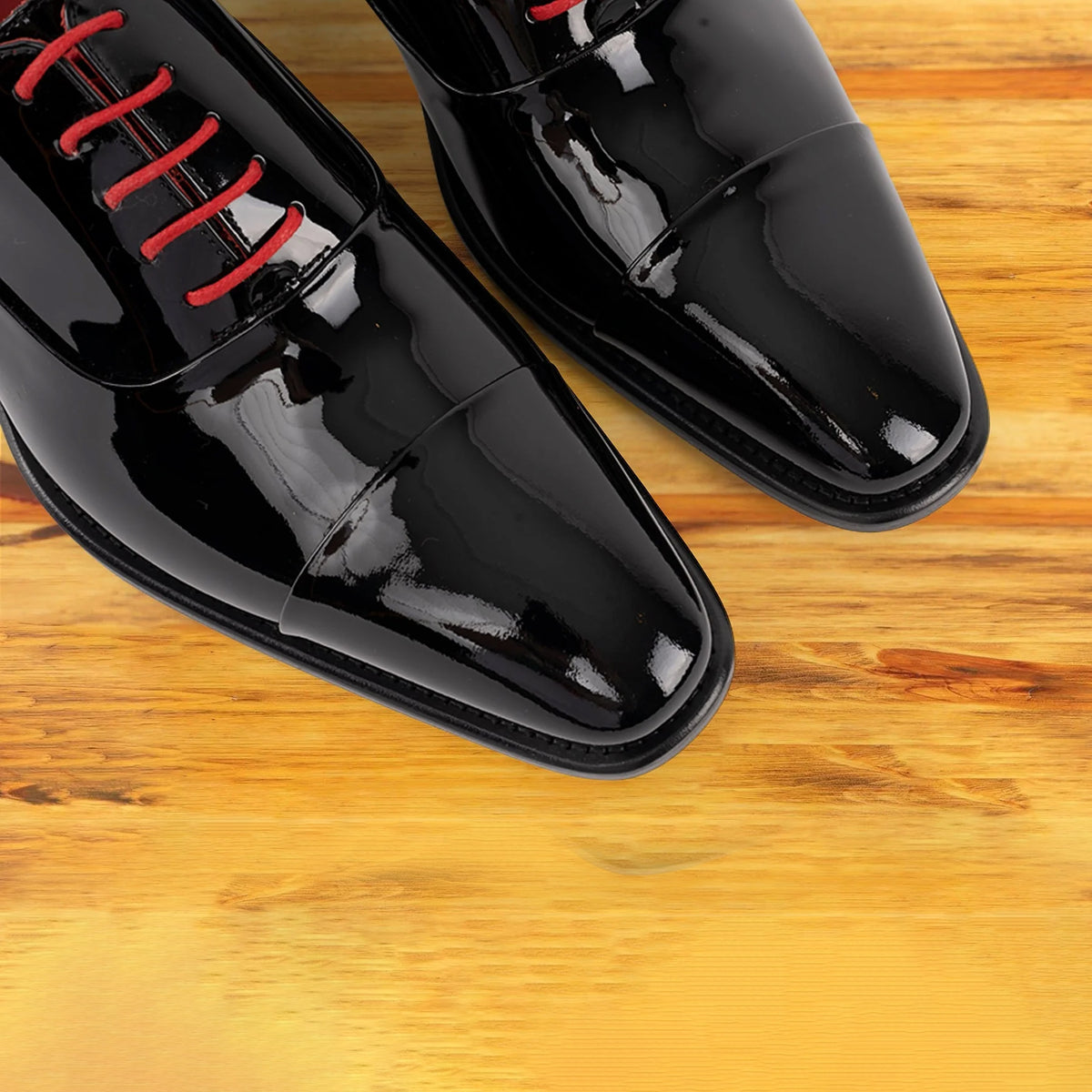 A close-up of a pair of glossy black patent leather dress shoes with bright red laces, resting on a warm wooden surface.
