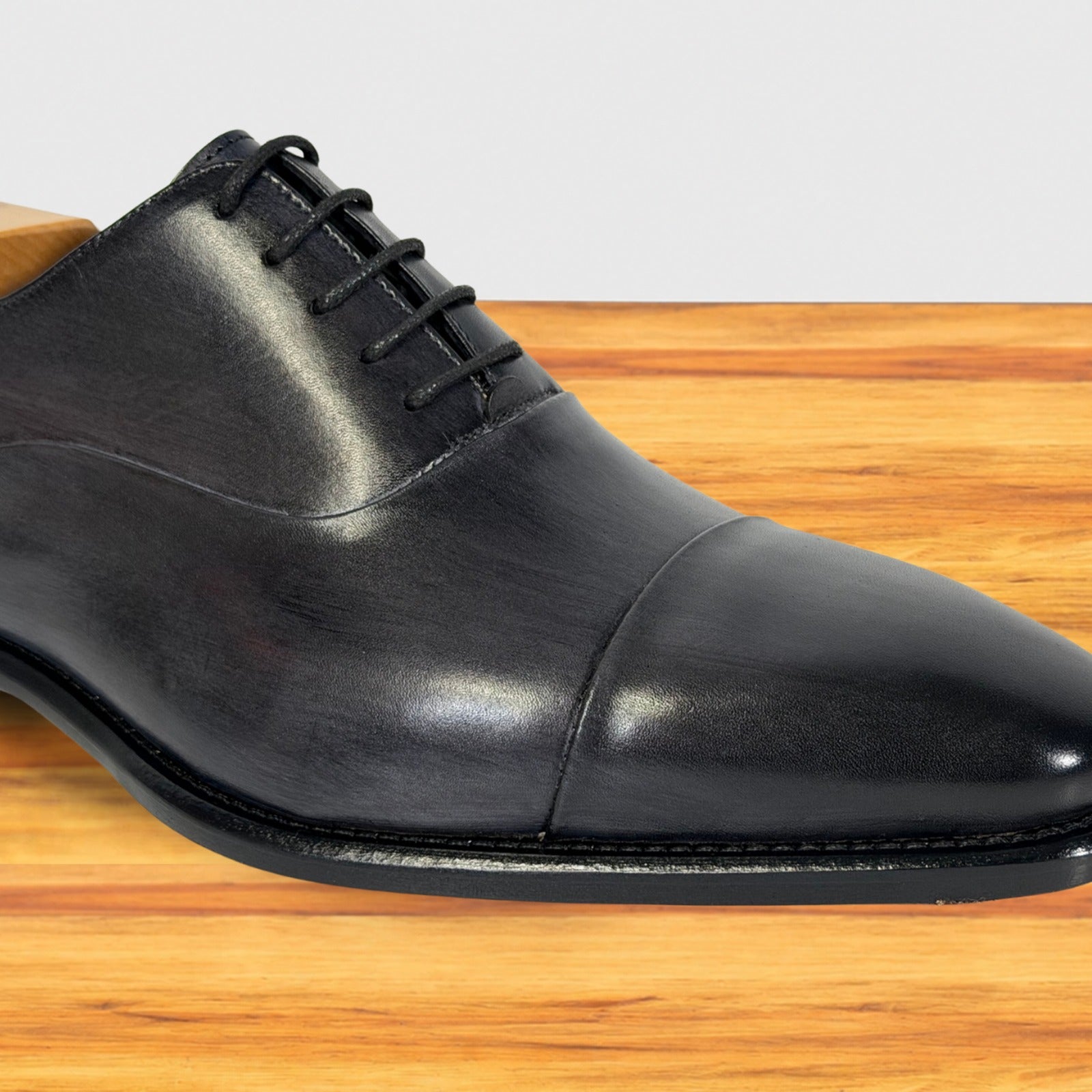 A gray leather cap-toe oxford dress shoe with laces, shown from a side angle on a wooden surface against a light gray background.