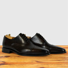A pair of Dark Brown leather cap-toe oxford shoes with black laces, displayed on a wooden surface against a light gray background.