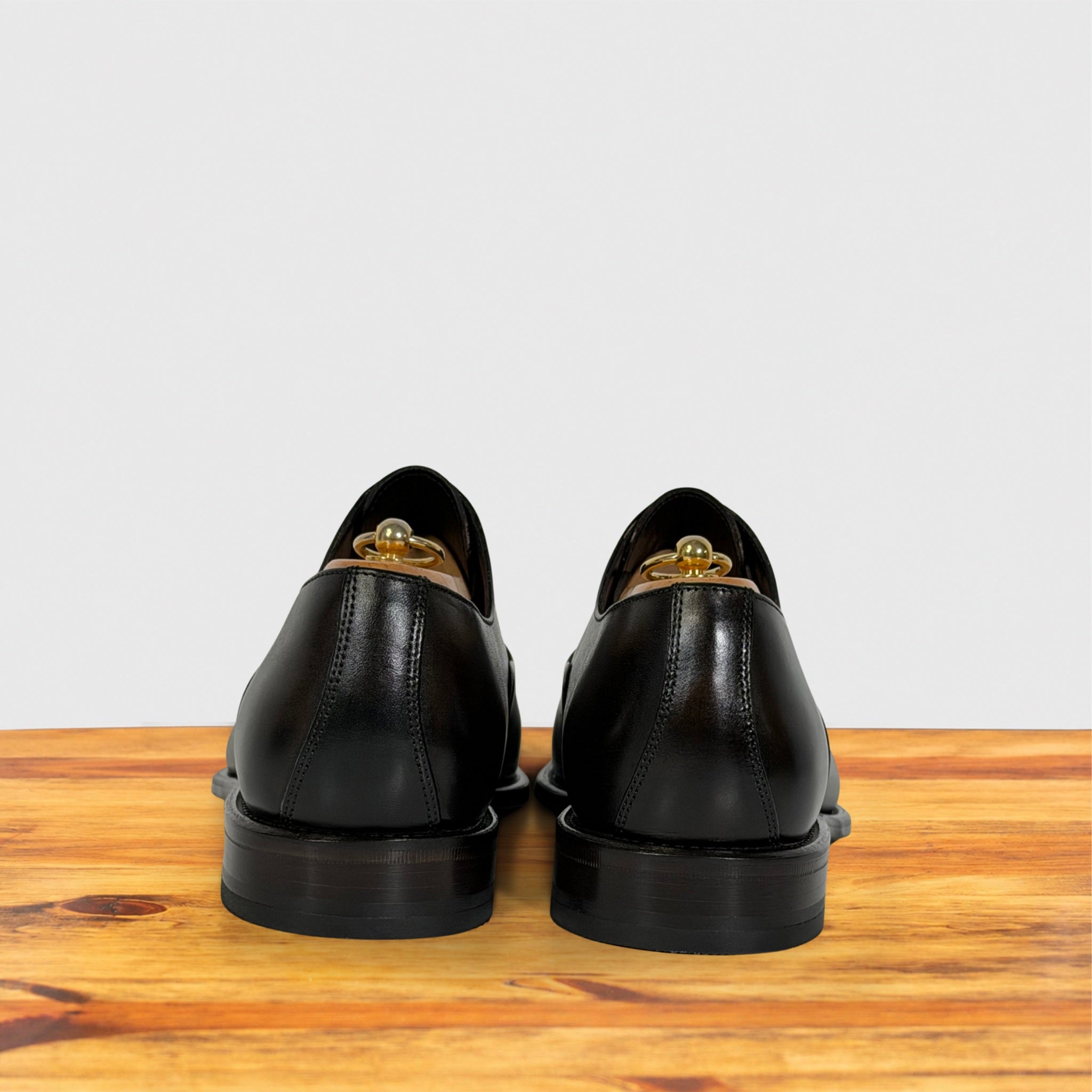 A pair of Dark Brown leather dress shoes shown from the back, resting on a wooden surface against a light gray background.