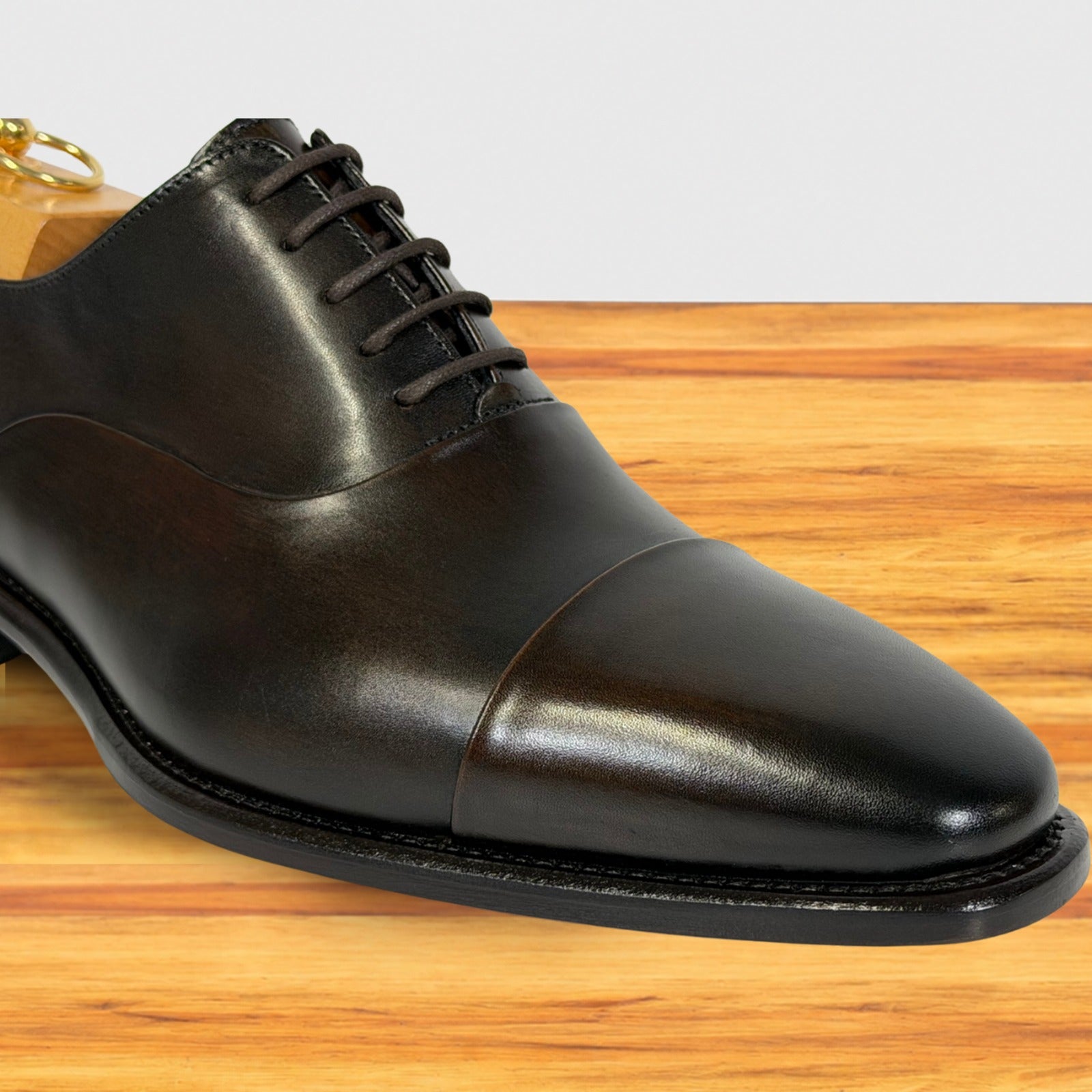 A close-up of a dark brown leather cap-toe oxford shoe resting on a wooden surface against a light gray background.