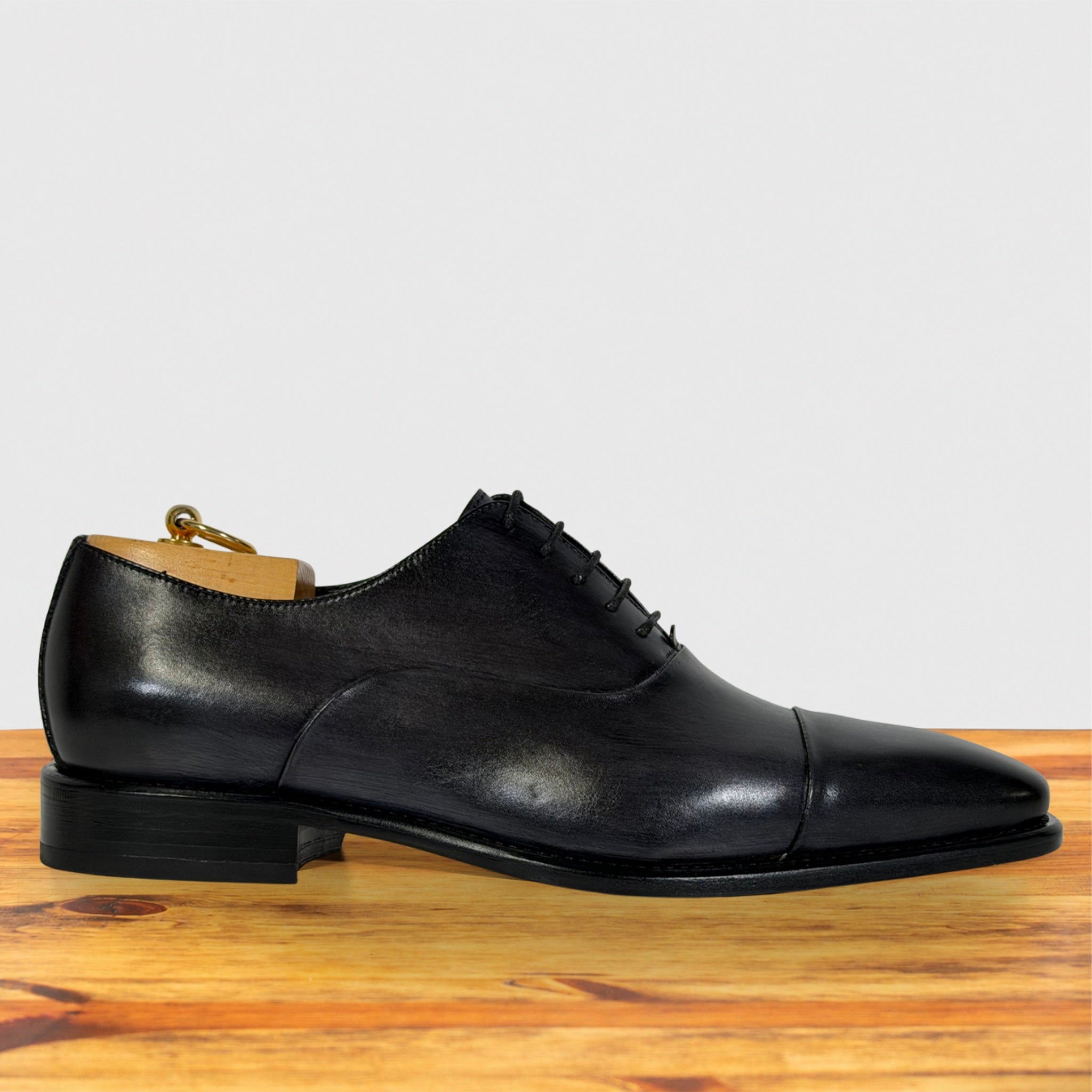 A Gray leather lace-up dress shoe with a cap toe, displayed on a wooden surface against a light gray background.