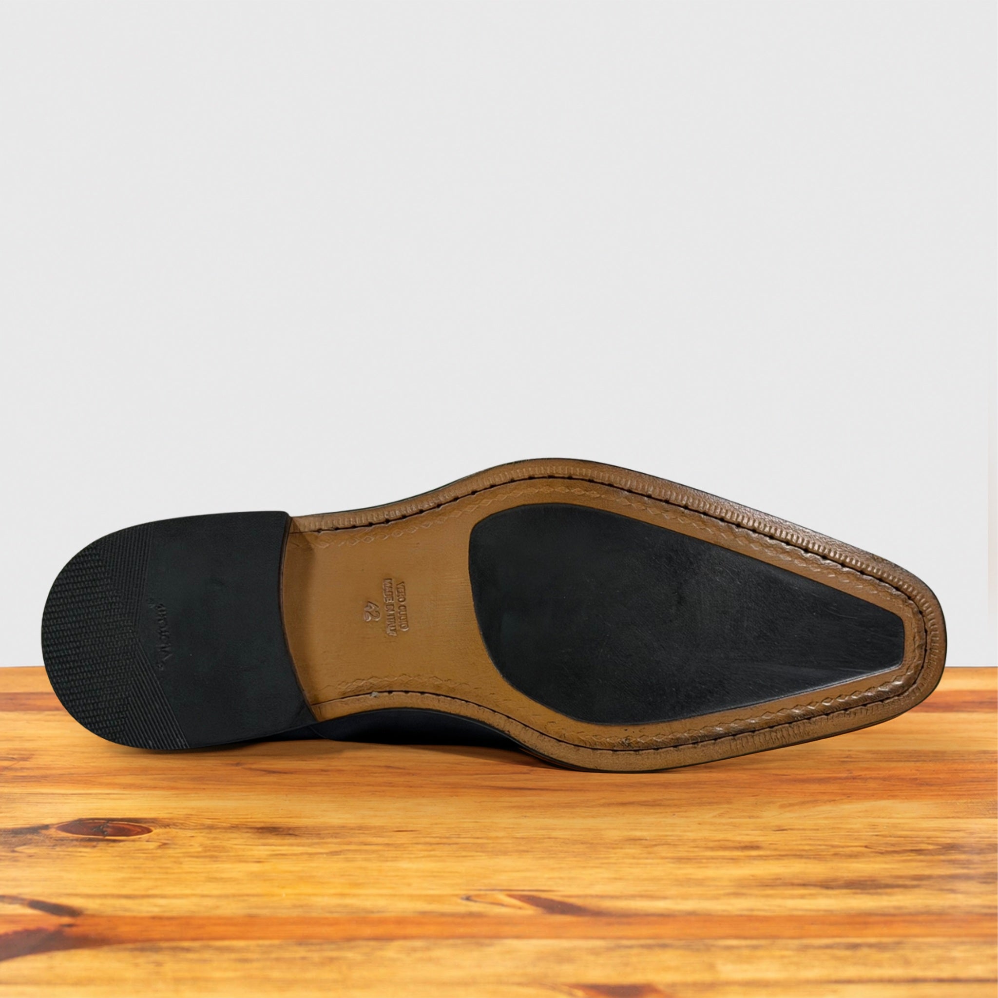 The outsole of a men's dress shoe, showing a black rubber tread, a tan leather midsole, and a black heel cap, displayed on a wooden surface against a light gray background.