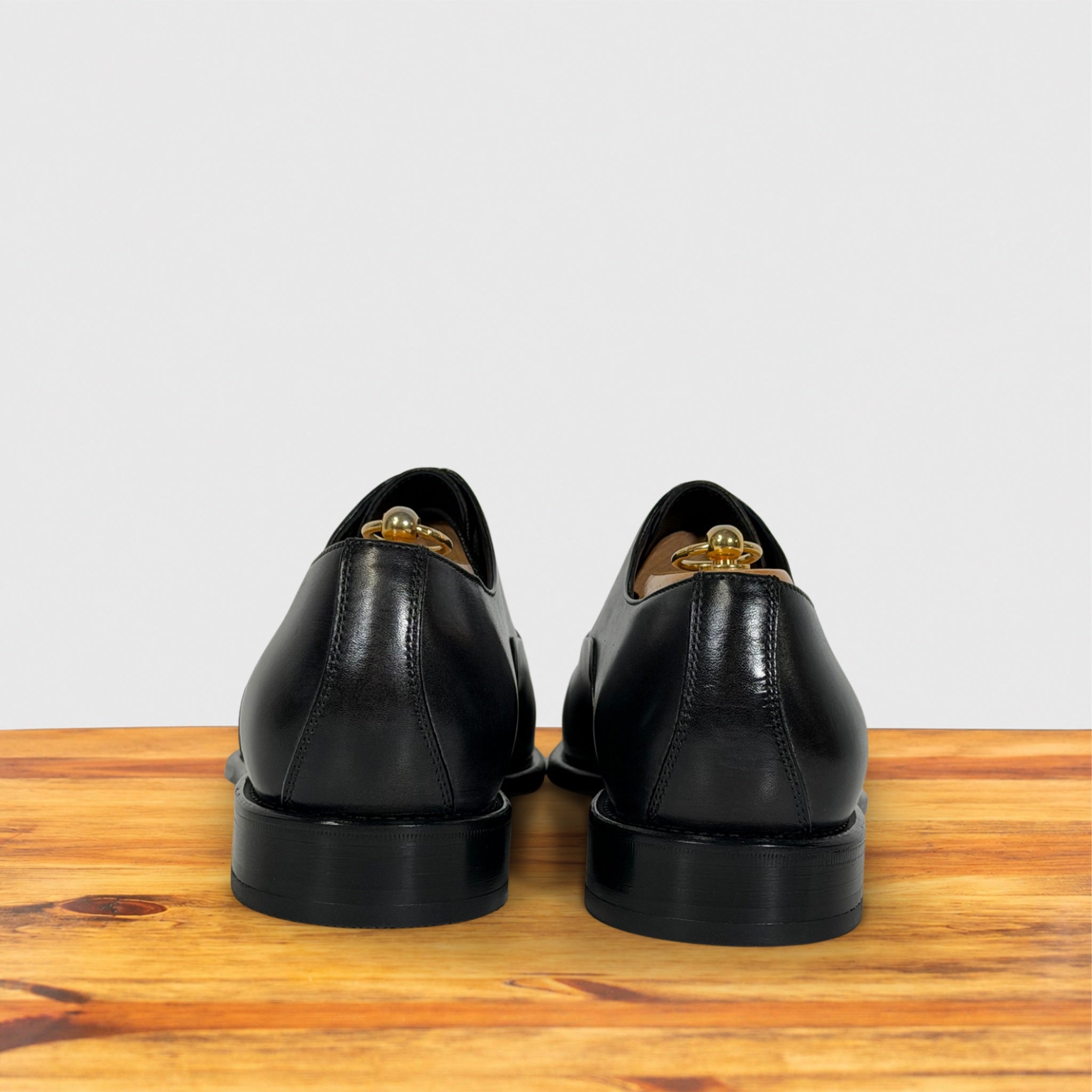 A pair of Gray leather dress shoes shown from the back, resting on a wooden surface against a light gray background.